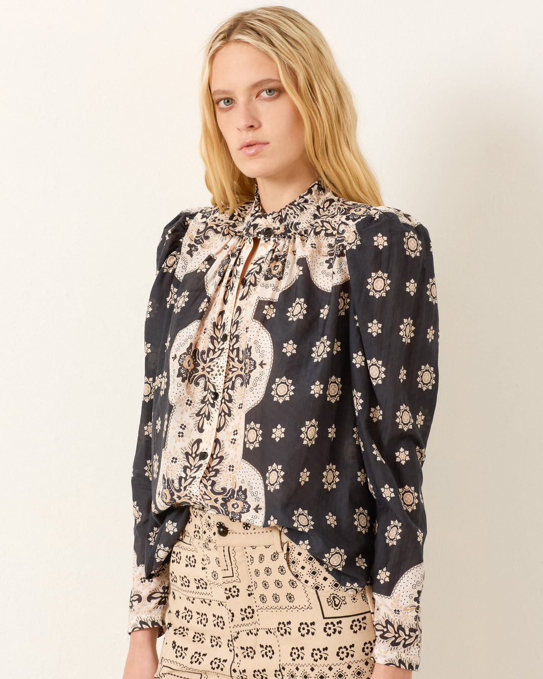 A woman with long blonde hair wears the Artemis Bronco Bandana Shirt, a patterned black and beige cotton-silk blend top with a bow tie at the neck, paired with a beige skirt featuring black geometric designs.