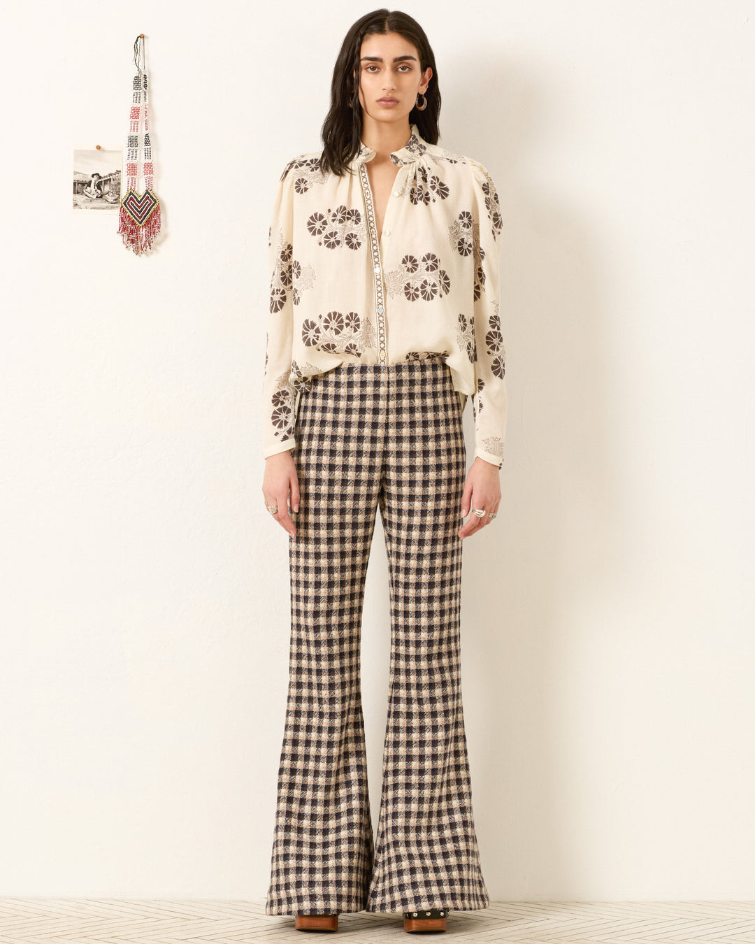 A person stands against a white wall wearing the Artemis Java Flower Shirt with dark floral patterns and high-waisted, flared checkered pants. Small wall decor is visible to the left.