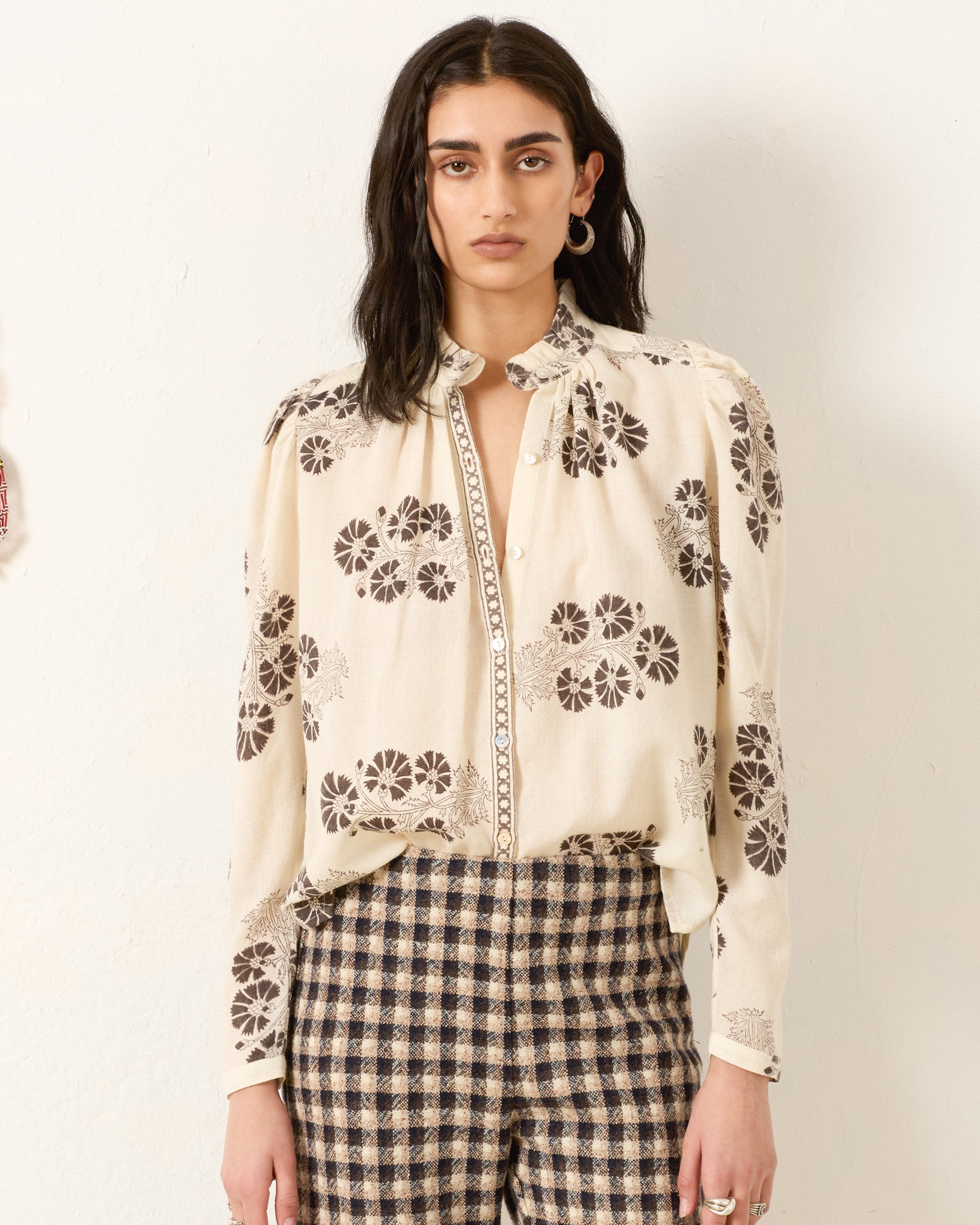 Wearing the Artemis Java Flower Shirt, a vintage-inspired blouse with hand-block floral prints, and checkered high-waisted pants, a person stands against a plain light-colored wall.