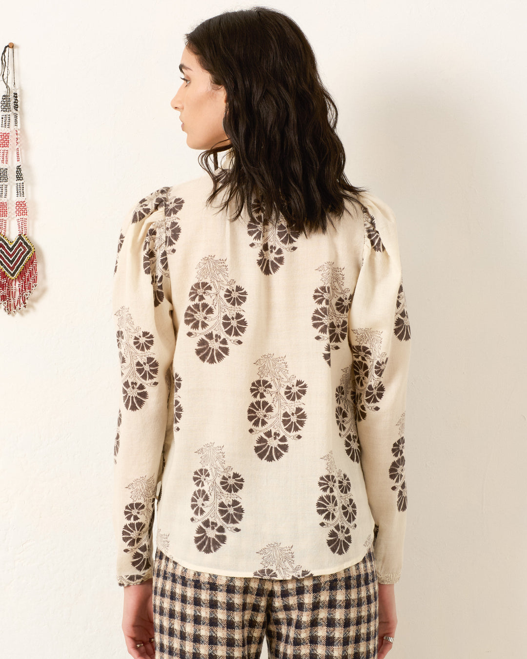 A woman, facing away, wears the Artemis Java Flower Shirt—a vintage-inspired cream blouse with brown floral prints—and checked pants, standing before a light-colored wall with a beaded hanging on the left.