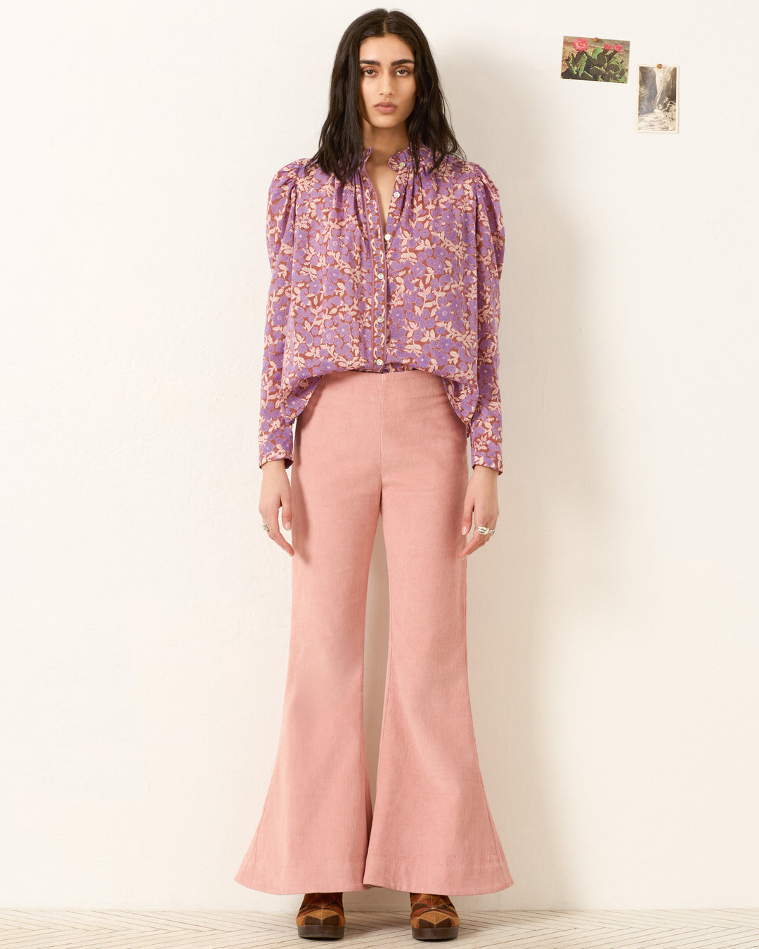 A woman stands against a plain wall wearing the Artemis Twilight Floral Shirt with puffed sleeves, paired with high-waisted pink flared pants. Two small photos hang on the wall behind her.