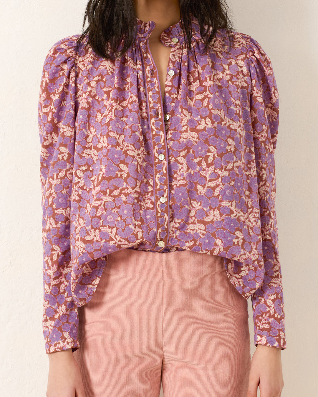 Wearing the Artemis Twilight Floral Shirt with a purple floral motif and pink high-waisted pants, the person stands against a plain off-white wall.