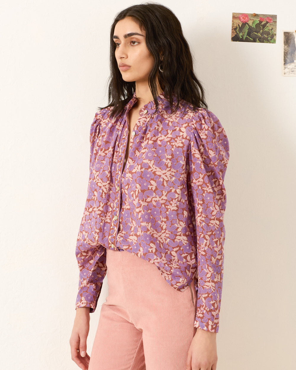 A woman stands against a light-colored wall wearing the Artemis Twilight Floral Shirt with a purple floral print and puffed sleeves, paired with high-waisted pink pants. Two small pictures hang on the wall behind her.