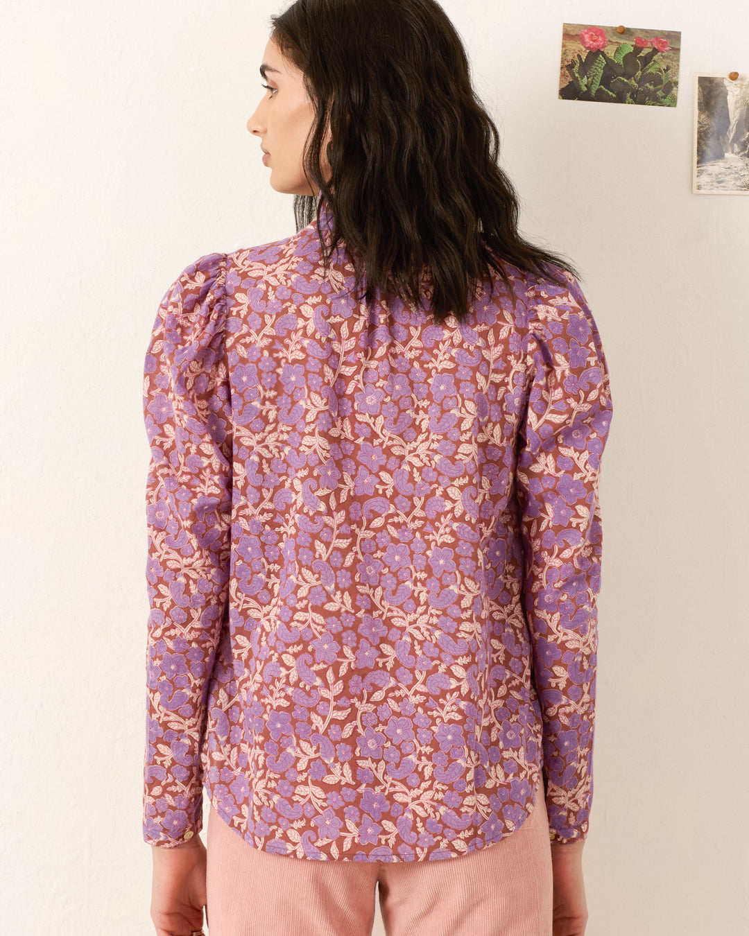 A woman with dark wavy hair faces away, wearing the Artemis Twilight Floral Shirt—a long-sleeve purple and pink floral blouse—and light pink pants. Two small pictures hang on the off-white wall.