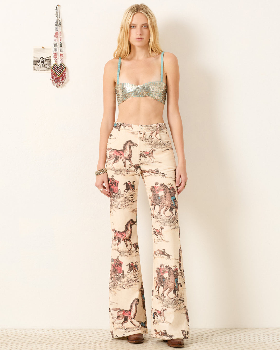 A woman stands against a plain wall wearing a metallic bralette and Bunny Canyon Cowboy Jean, which features high-waisted, flared legs with a vintage horse and carriage cowboy print.