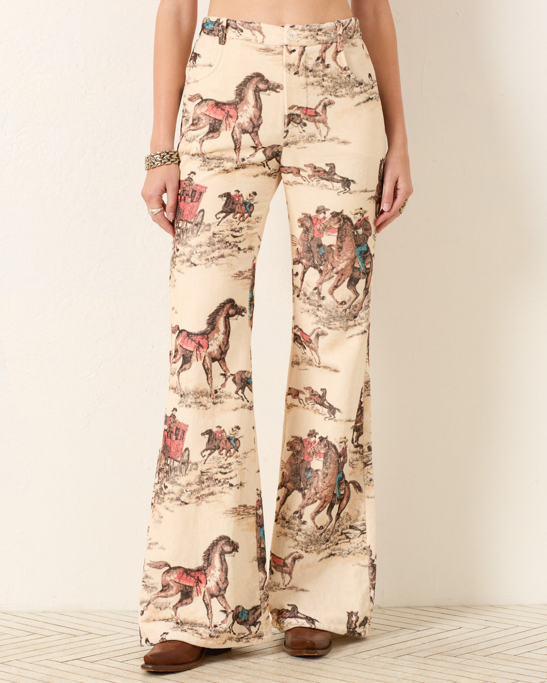 A woman models the Bunny Canyon Cowboy Jean—high-waisted, flared pants with a beige base and cowboy print in brown, red, and blue horse-and-rider motifs. Brown shoes complete the look.