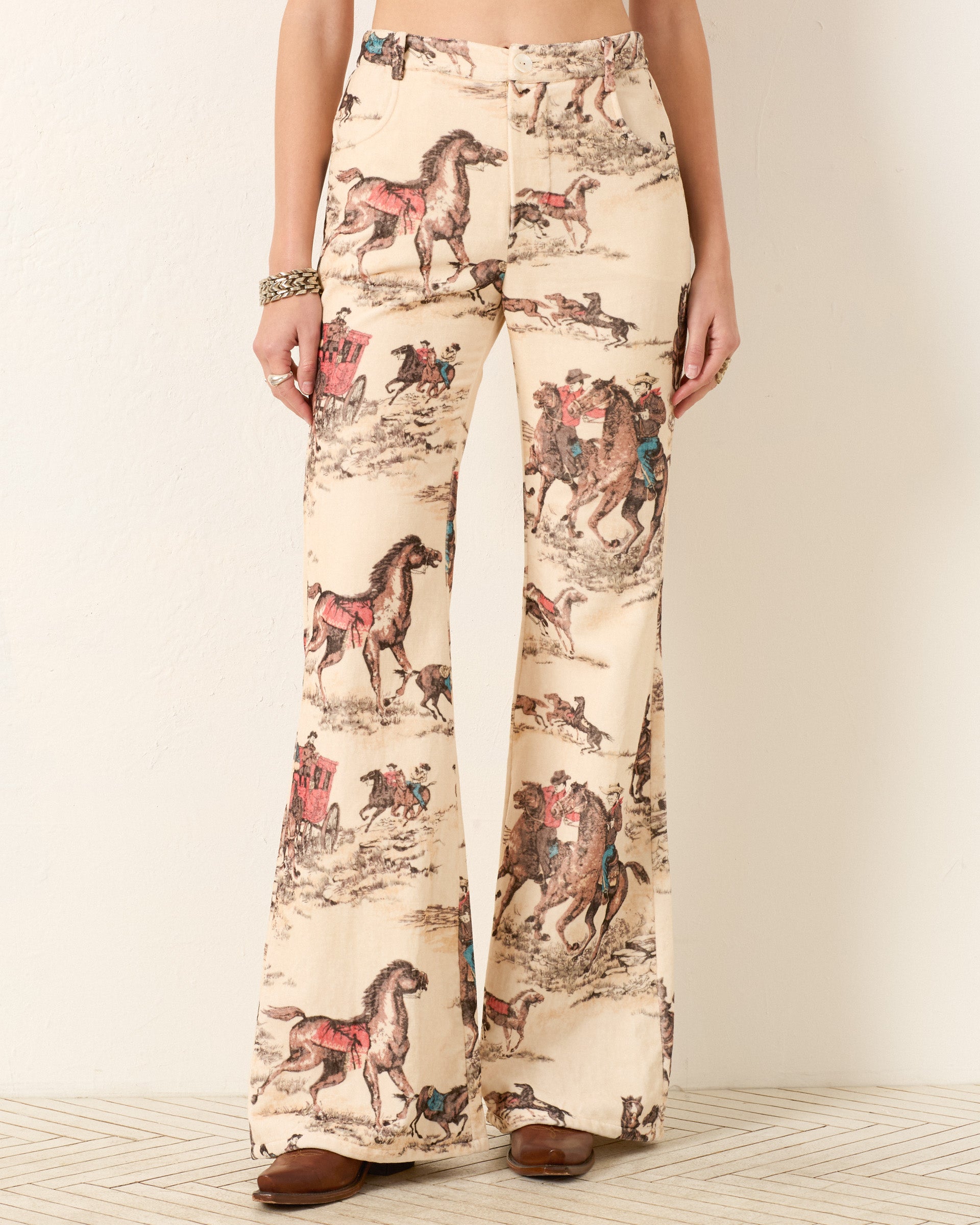 A woman models the Bunny Canyon Cowboy Jean—high-waisted, flared pants with a beige base and cowboy print in brown, red, and blue horse-and-rider motifs. Brown shoes complete the look.