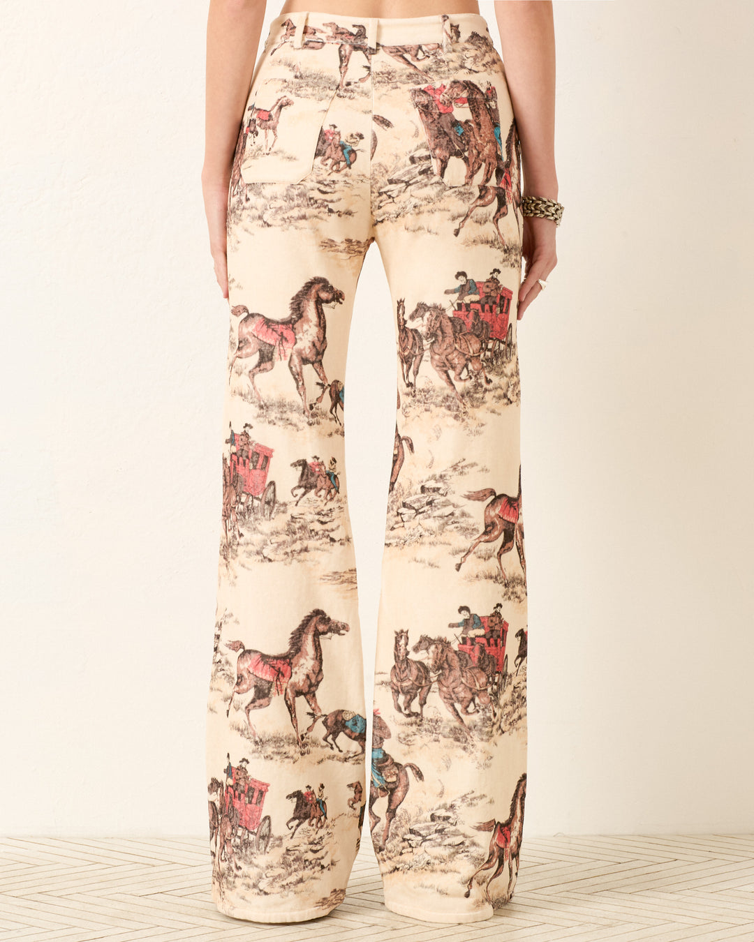 Model wears Bunny Canyon Cowboy Jean—high-waisted, wide-leg cotton-velvet pants with a beige base and illustrated cowboy print showing horses, riders, and stagecoaches.