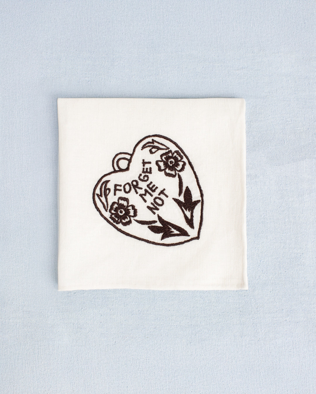 The Cai Java Heart Handkerchief, made of white linen-cotton and featuring a black embroidered heart, floral design, and “FORGET ME NOT” text, is displayed on a light blue background.