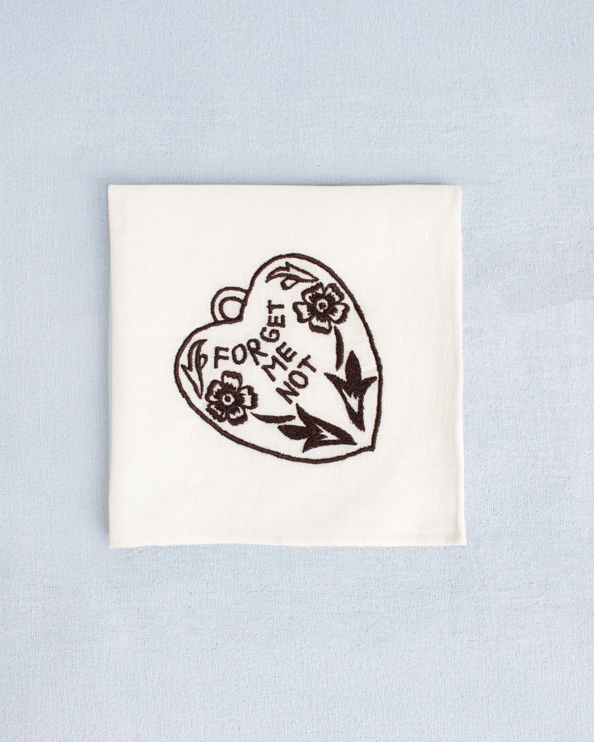 The Cai Java Heart Handkerchief, made of white linen-cotton and featuring a black embroidered heart, floral design, and “FORGET ME NOT” text, is displayed on a light blue background.