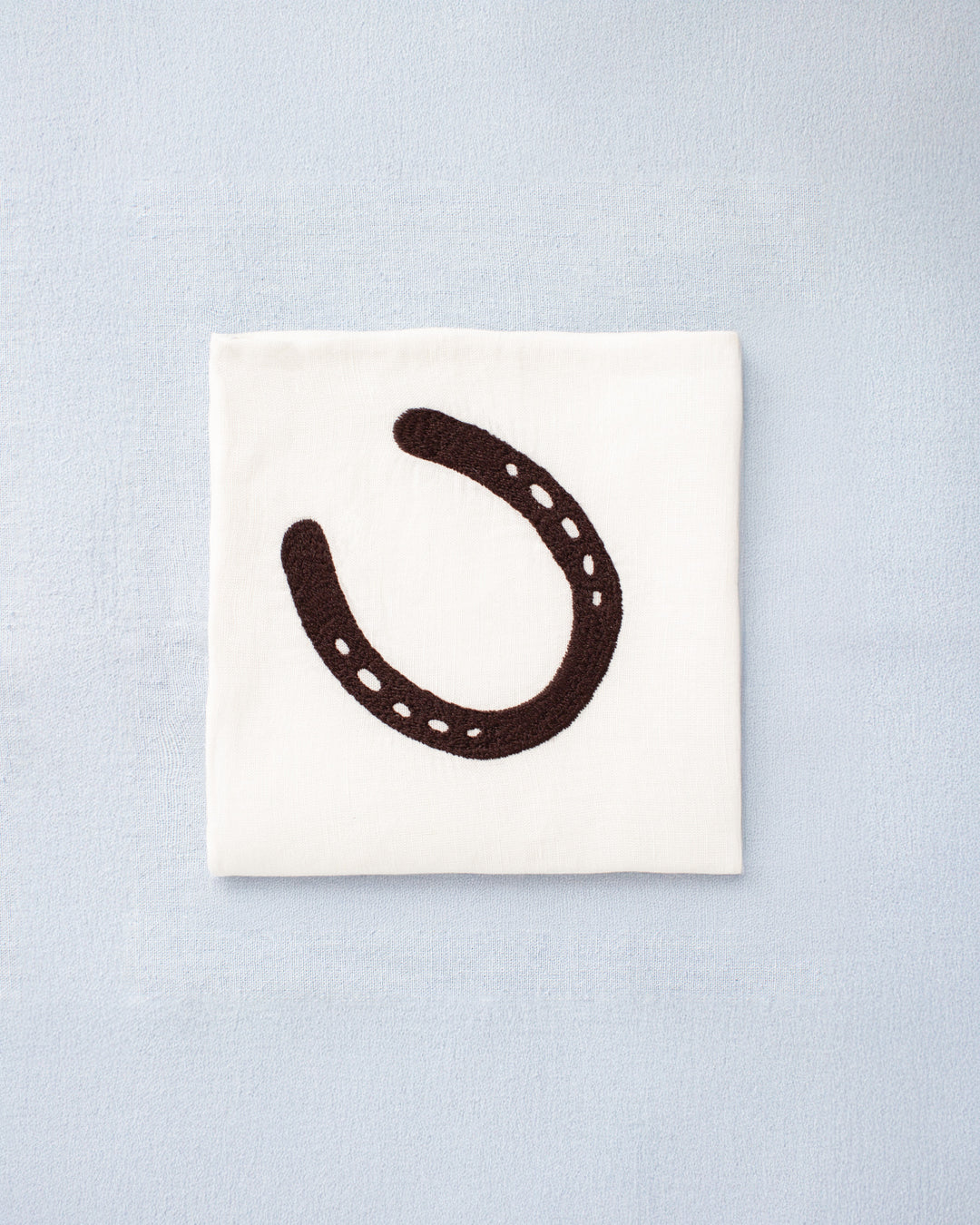The Cai Horseshoe Handkerchief, made from white linen-cotton and featuring a black horseshoe motif, rests on a light blue surface.