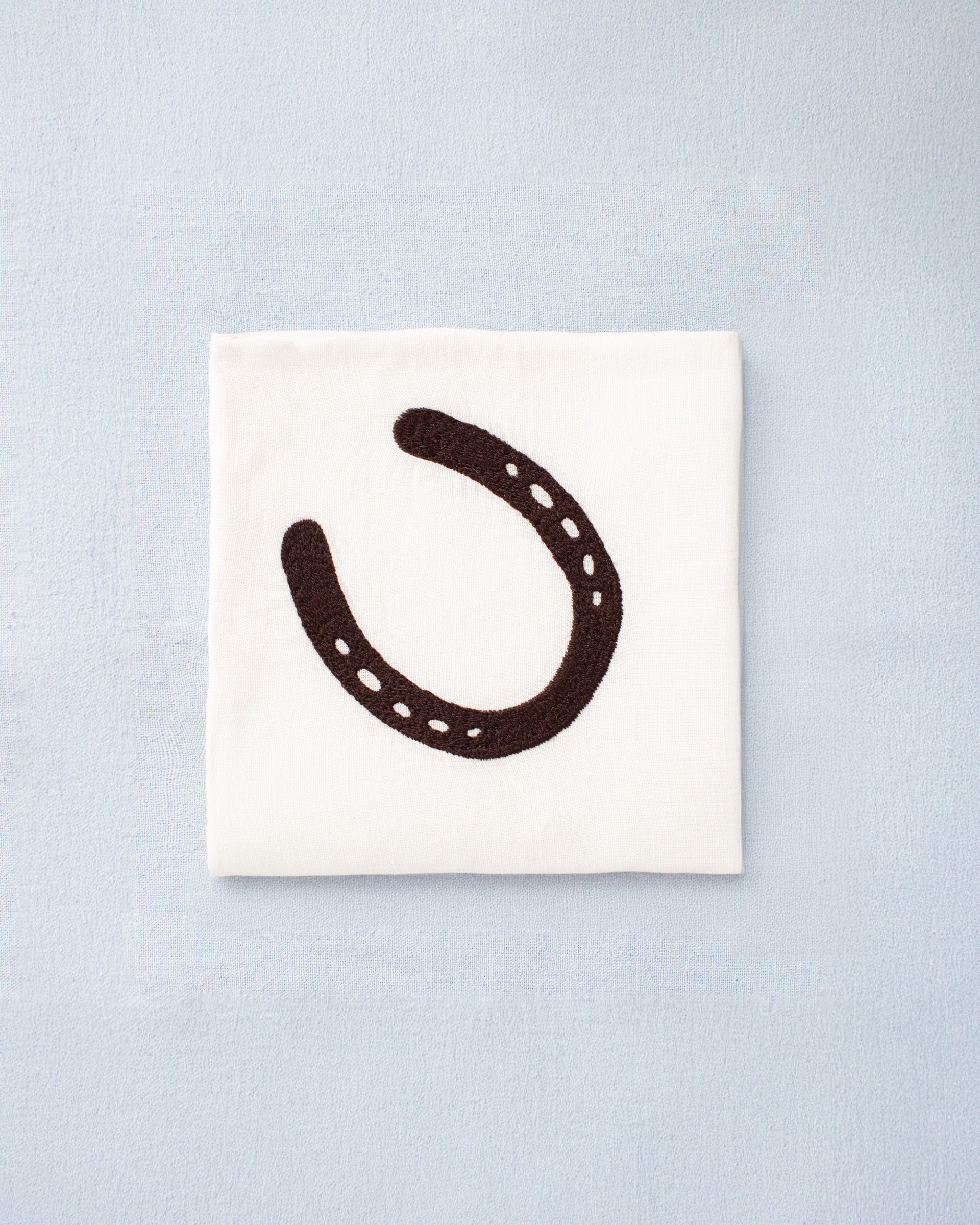 The Cai Horseshoe Handkerchief, made from white linen-cotton and featuring a black horseshoe motif, rests on a light blue surface.