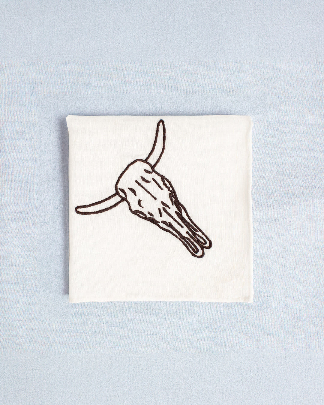 The Cai Cow Skull Handkerchief, made from a linen-cotton blend and featuring a black outline of a Western cow skull, is neatly folded on a light blue surface—perfect as a unique good luck gift.