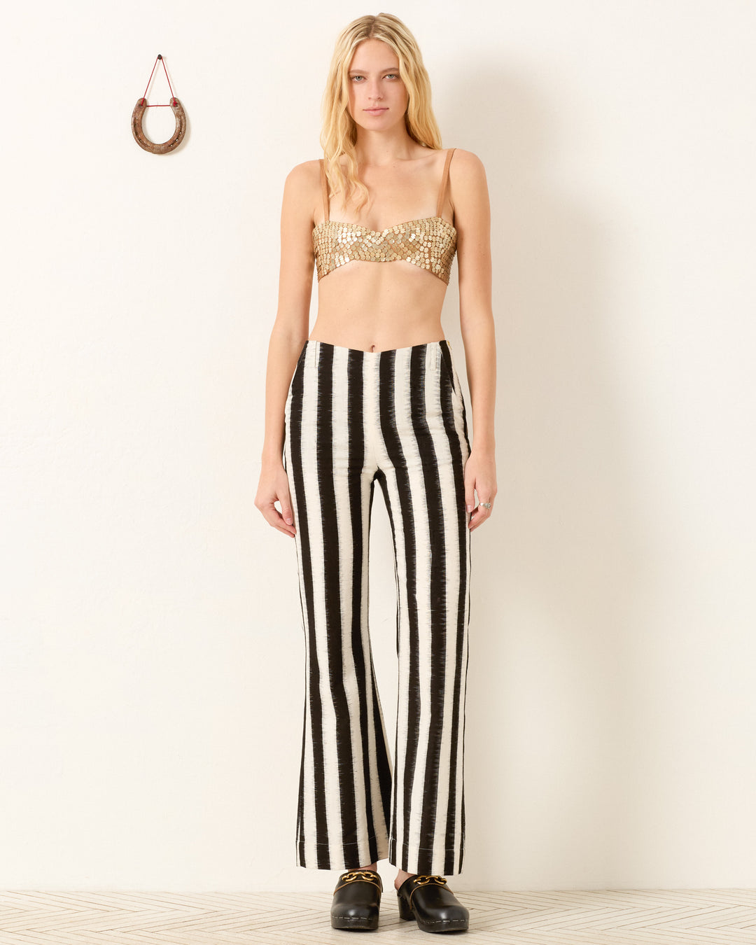 A woman stands indoors wearing a gold sequin bandeau top, Charlie Coal Stripe Pant, and black shoes. A horseshoe hangs on the wall behind her.