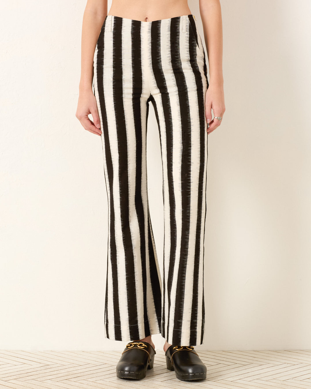 Person wearing the Charlie Coal Stripe Pant, a high-waisted black and white vertically striped flared leg pant, paired with black slip-on shoes, standing against a plain light wall.