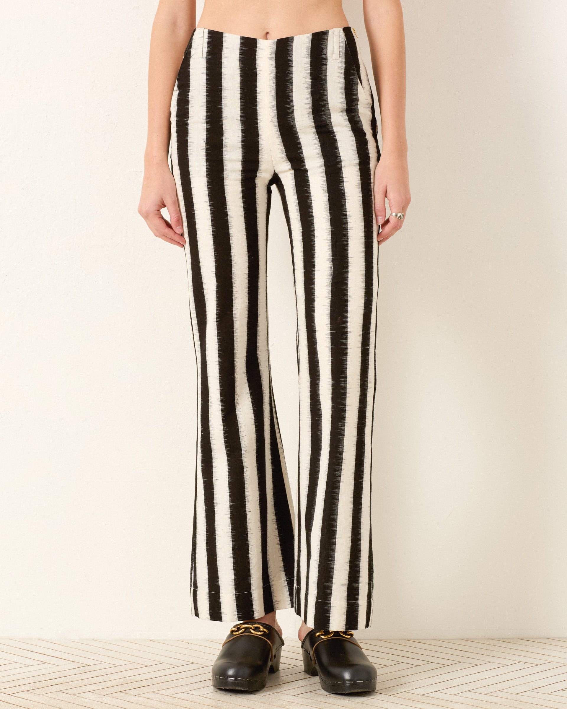 Person wearing the Charlie Coal Stripe Pant, a high-waisted black and white vertically striped flared leg pant, paired with black slip-on shoes, standing against a plain light wall.