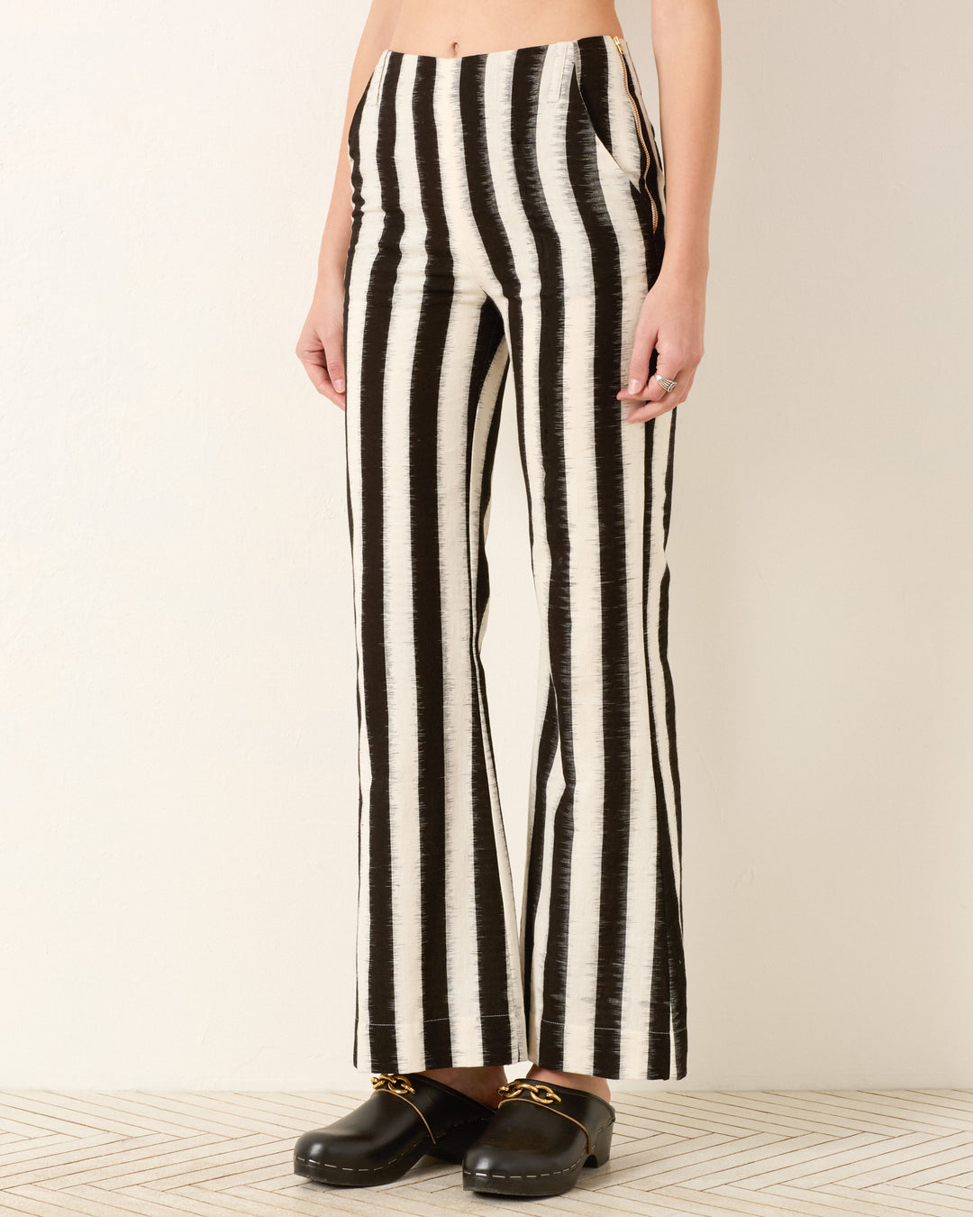 Person wearing the Charlie Coal Stripe Pant, featuring black and white vertical stripes with a flared leg, paired with black slip-on shoes with gold chain detail, standing against a plain light-colored wall.