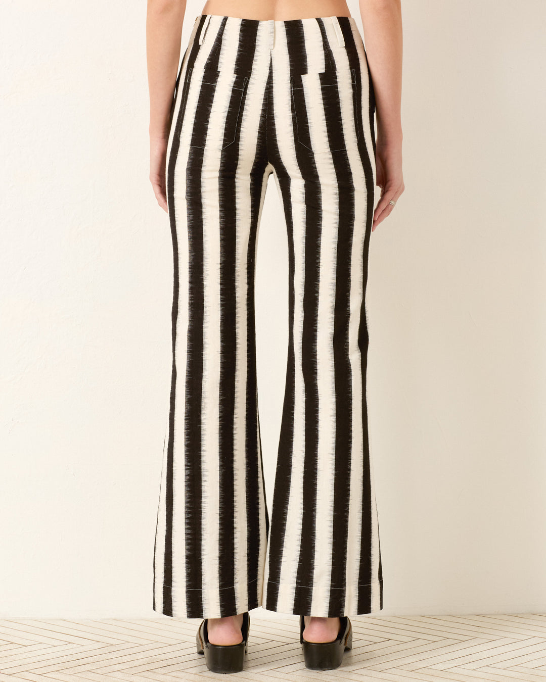 A person shown from the back wears the Charlie Coal Stripe Pant—black and white vertically striped flared-leg pants crafted from ikat cotton—with black platform shoes.