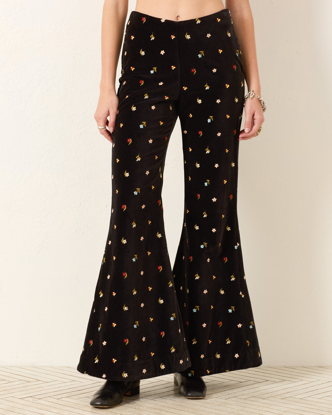 Wearing the Charlie Onyx Bouquet Pant—high-waisted, black cotton-velvet with a bell-bottom flare and small multicolor floral print—paired with black shoes and a bracelet.