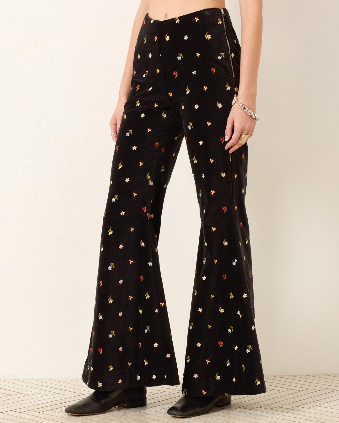 Wearing the Charlie Onyx Bouquet Pant—black cotton-velvet pants with colorful floral embroidery and a bell-bottom flare—paired with black shoes and a silver bracelet, standing against a neutral background.