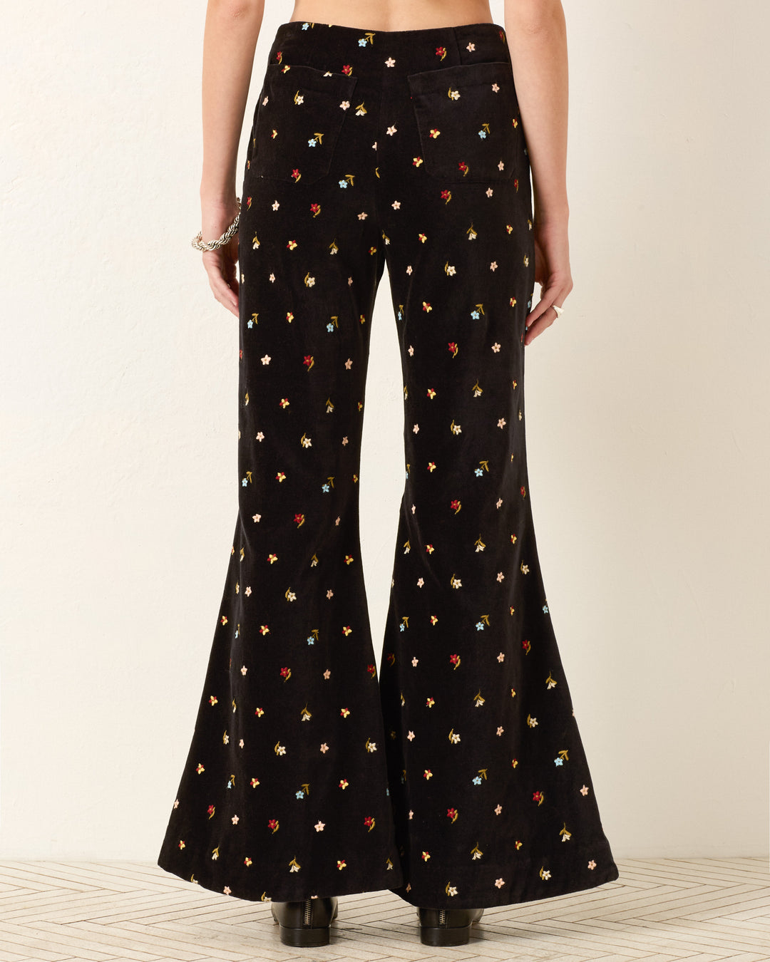 Someone is seen from the back wearing the Charlie Onyx Bouquet Pant—high-waisted, black cotton-velvet pants with a bell-bottom flare and a vibrant small floral print.