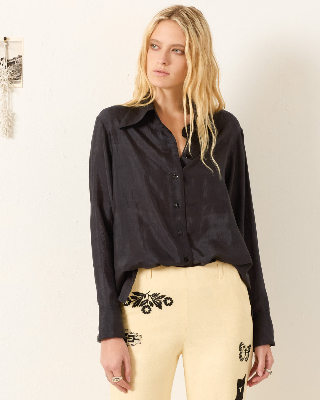 A woman with long blonde hair wears the Dana Onyx Silk Shirt and high-waisted beige pants with black embroidery, standing against a plain light-colored wall.