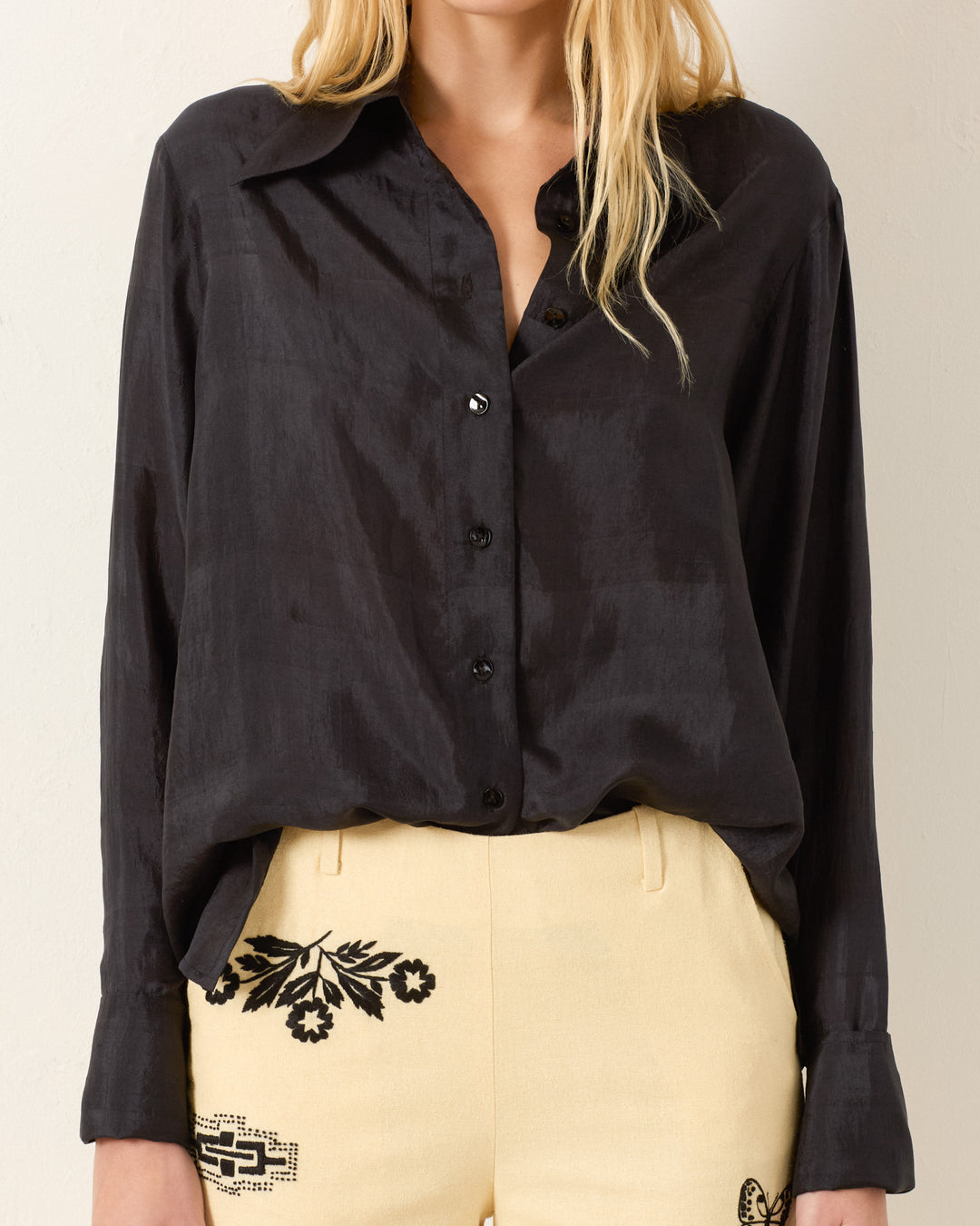 A woman wears the Dana Onyx Silk Shirt, a vintage-inspired black button-up with French cuffs, tucked into high-waisted cream pants featuring black embroidered designs.
