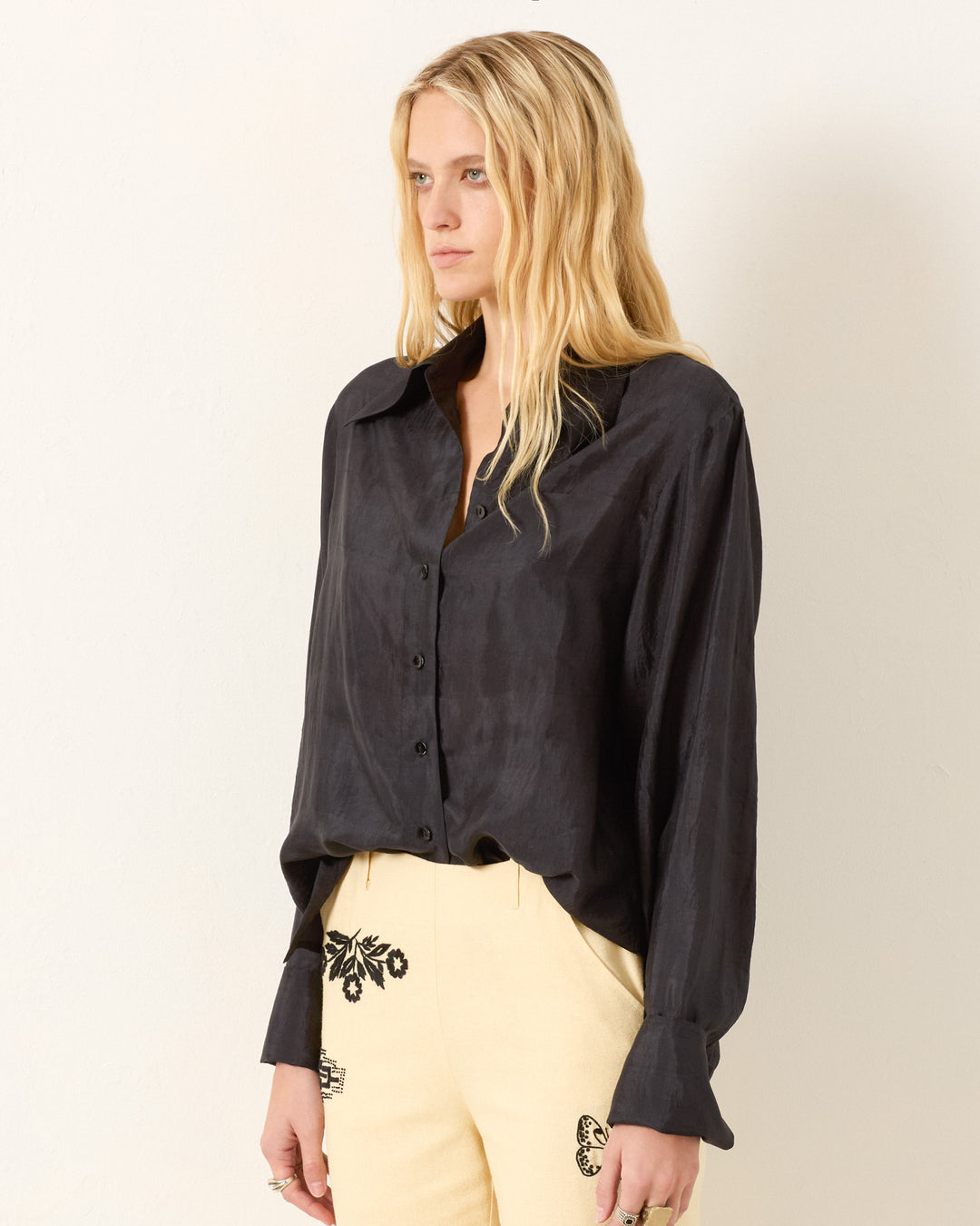 A woman with long blonde hair wears the Dana Onyx Silk Shirt, a vintage-inspired black button-up with French cuffs, paired with cream pants featuring black embroidery, standing against a plain light-colored wall.