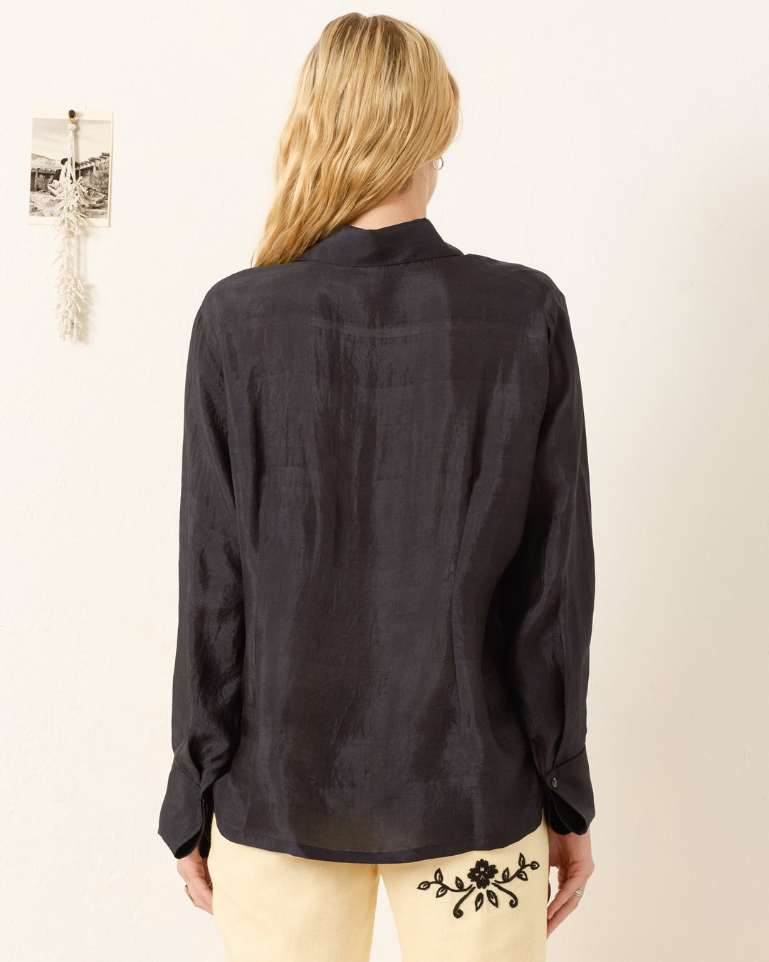 A person with long blonde hair is photographed from the back wearing the Dana Onyx Silk Shirt—a vintage-inspired black long-sleeve shirt with French cuffs—paired with cream pants featuring black embroidery.