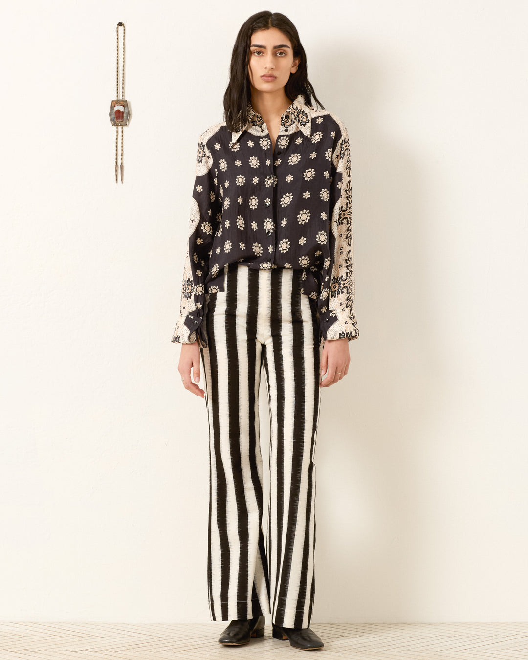 A woman stands against a white wall wearing the Dana Bronco Bandana Shirt with French cuffs and black-and-white vertical striped pants, paired with black shoes. A small wall clock hangs to her left.