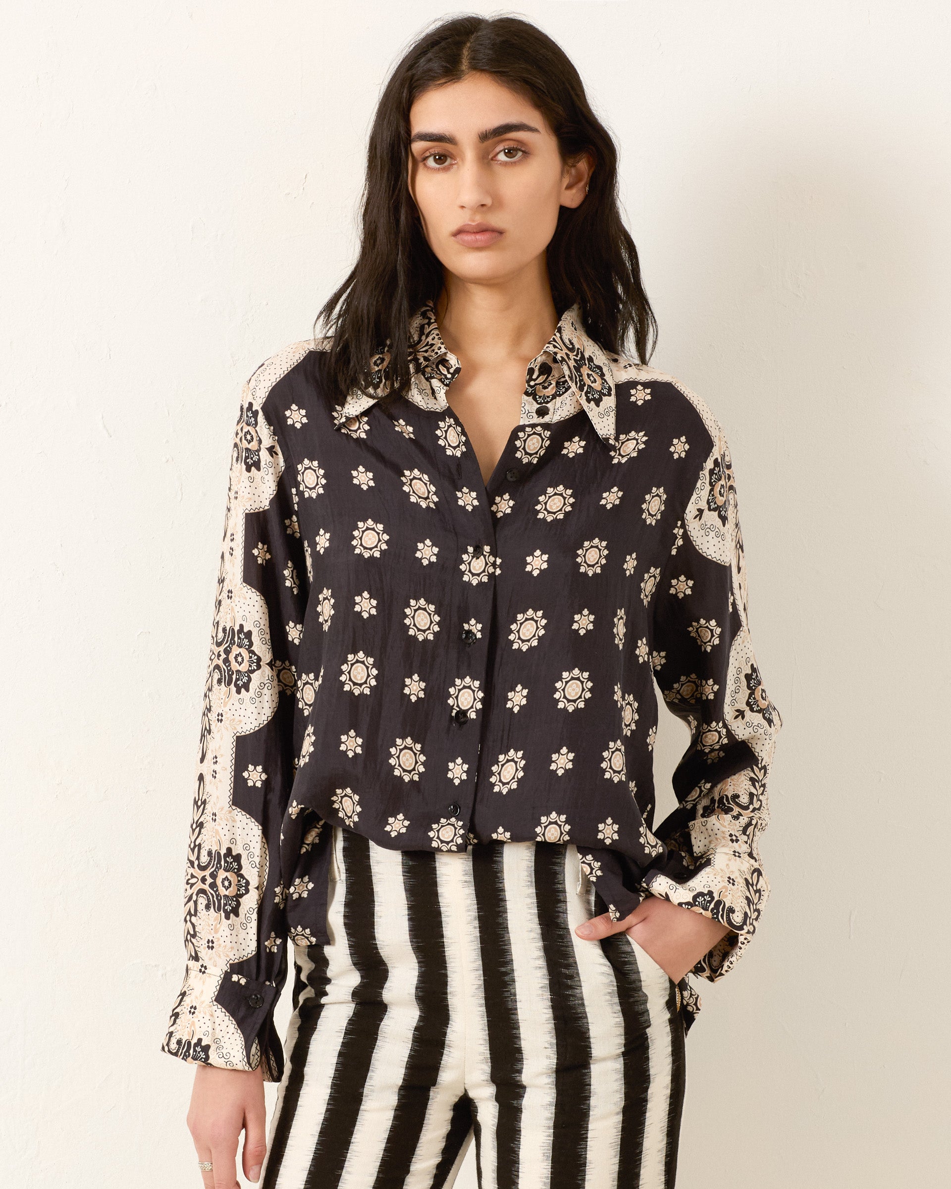 A person wearing the Dana Bronco Bandana Shirt, featuring a black and white bandana print with floral designs, and black and white vertically striped pants stands against a plain wall.