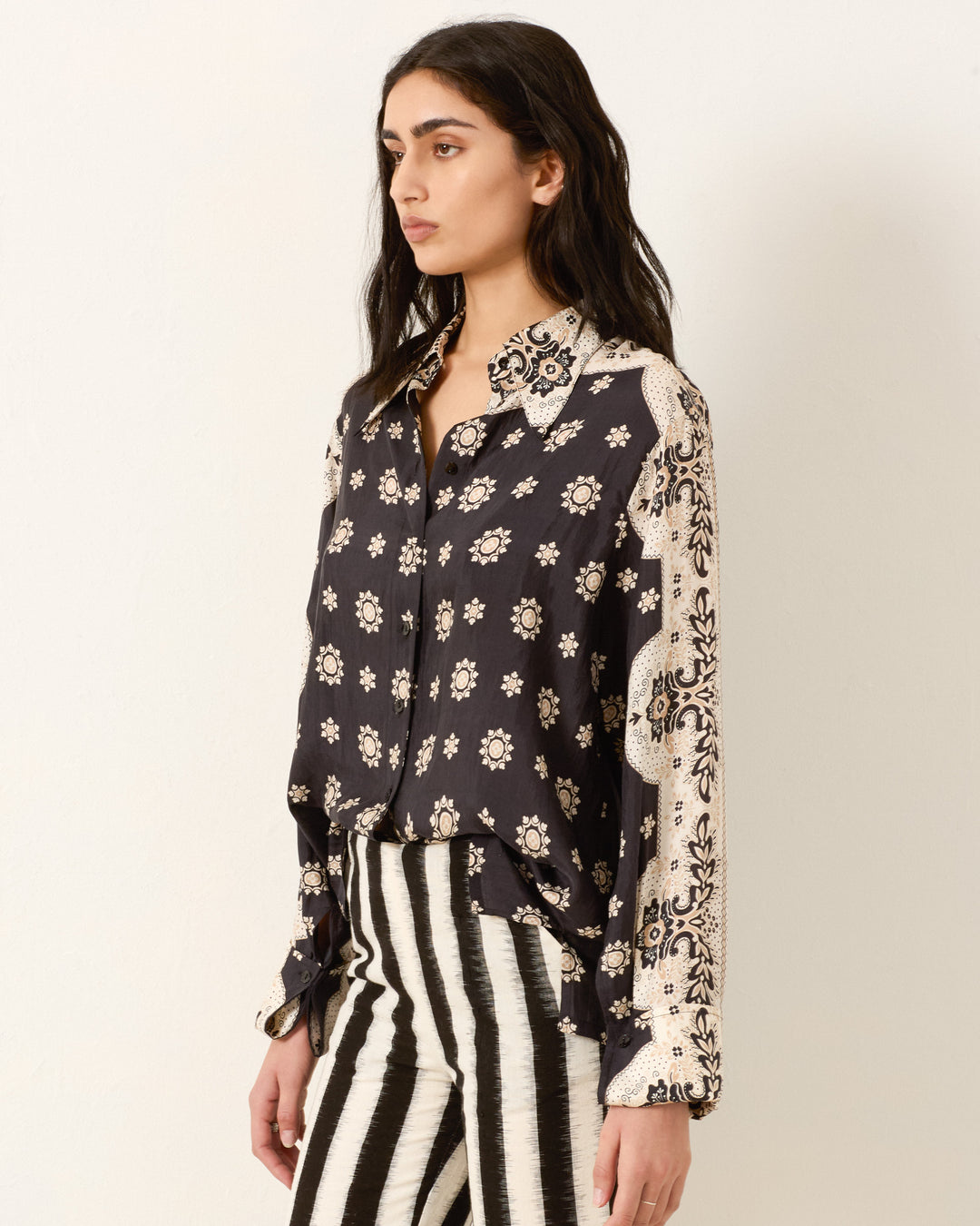 A woman stands wearing the Dana Bronco Bandana Shirt, featuring hand-loomed silk and patterned cream sleeves, paired with black-and-white vertical striped pants.