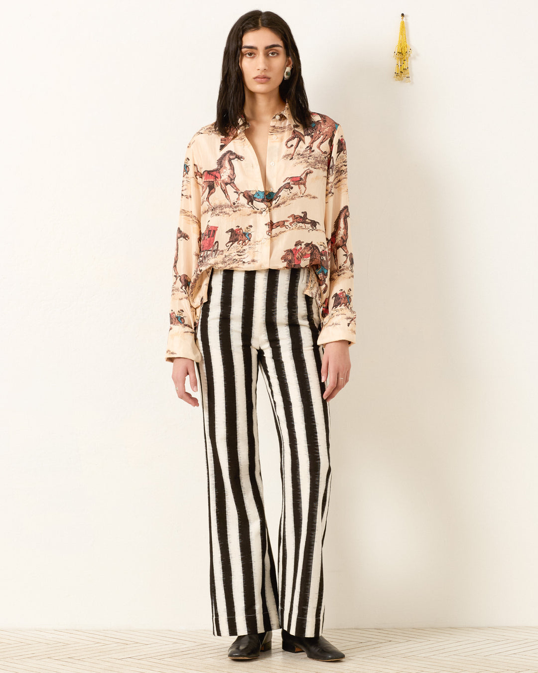 A person stands against a plain wall wearing the beige Dana Canyon Cowboy Silk Shirt with horse prints and black-and-white vertically striped wide-leg pants, paired with black shoes.