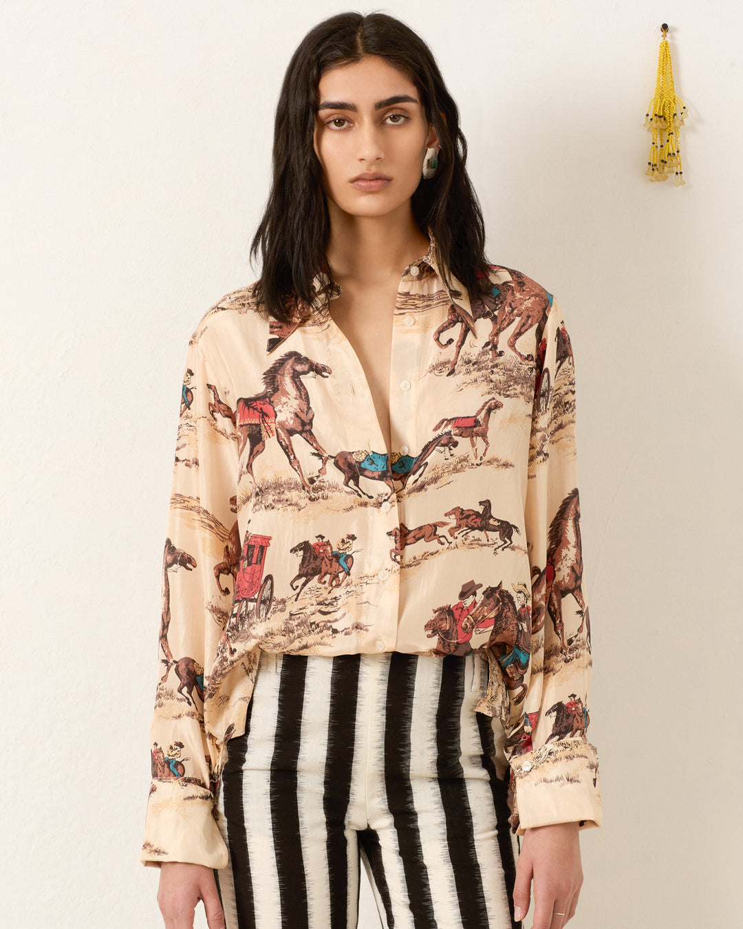 A woman with long dark hair wears the Dana Canyon Cowboy Silk Shirt, featuring a horse print, paired with black-and-white striped pants. She stands against a plain wall accented by a hanging yellow tassel.
