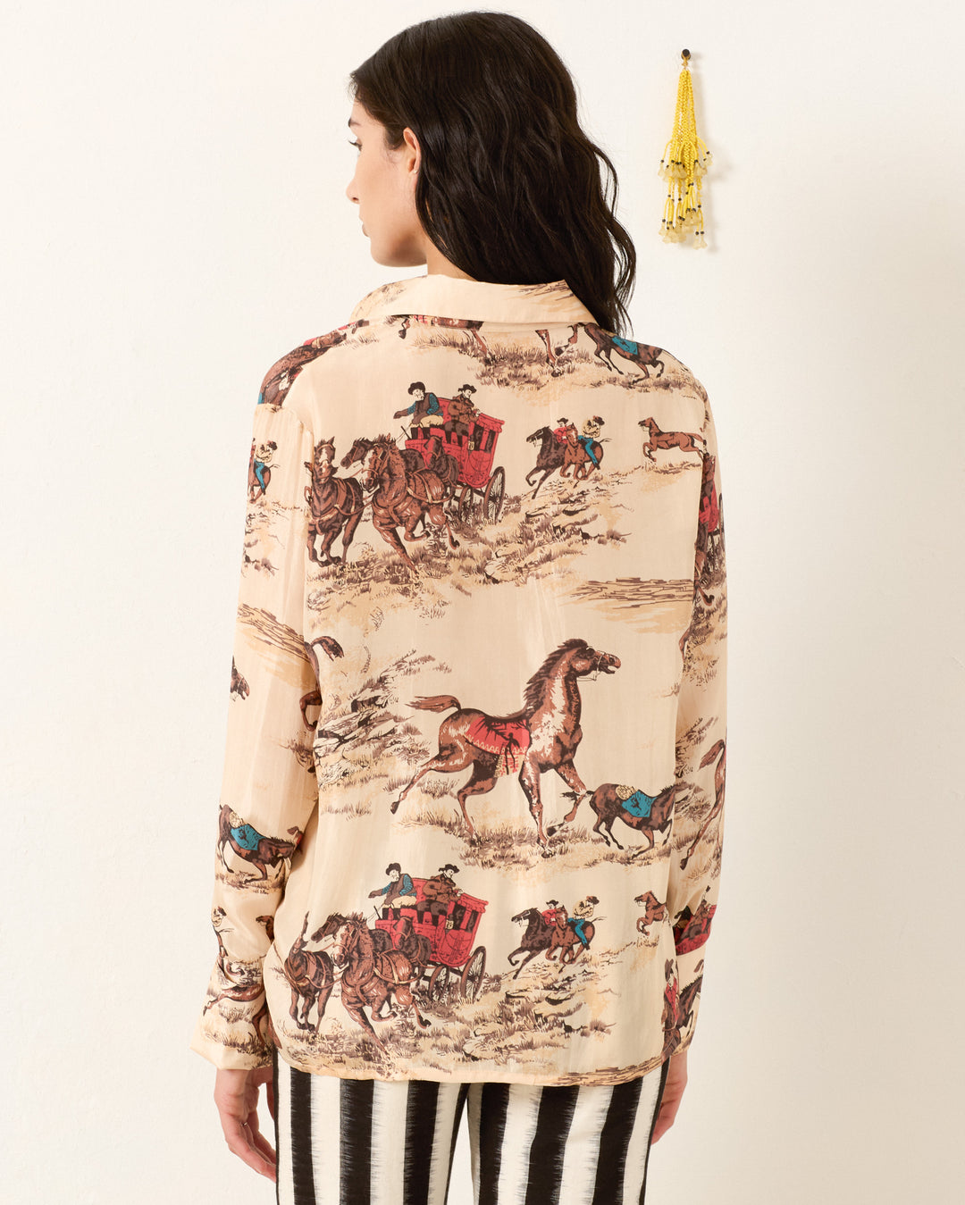 A woman faces away from the camera wearing the Dana Canyon Cowboy Silk Shirt in beige, featuring a horse and carriage print, paired with black and white striped pants.