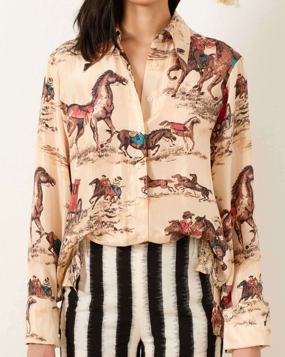 A woman wears the Dana Canyon Cowboy Silk Shirt in beige with a horse and cowboy print, styled with high-waisted black and white striped pants.