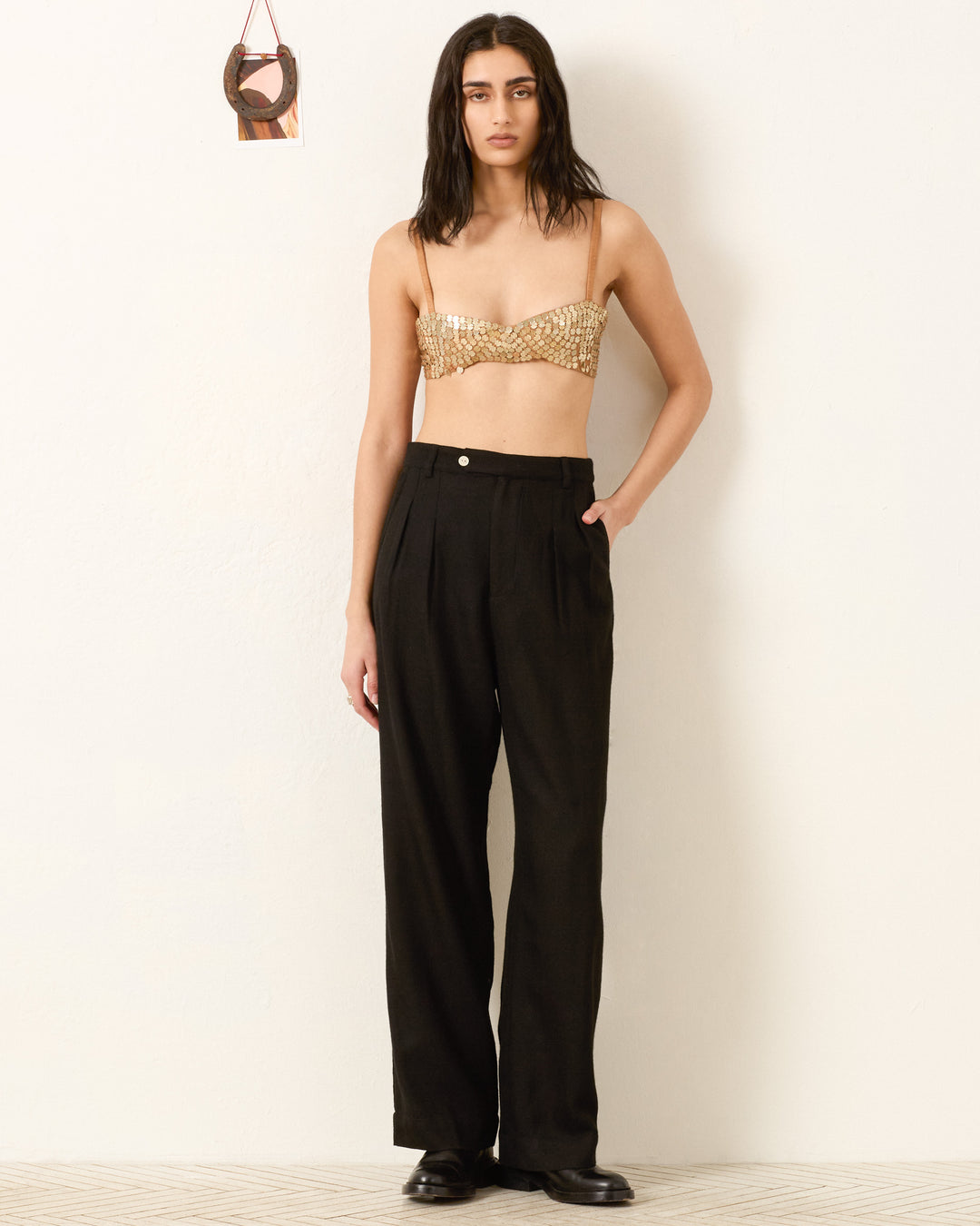 A woman stands against a white wall wearing a gold sequin bralette, the Diana Noir Wool Pant inspired by menswear tailoring, and black shoes, with one hand in her pocket.