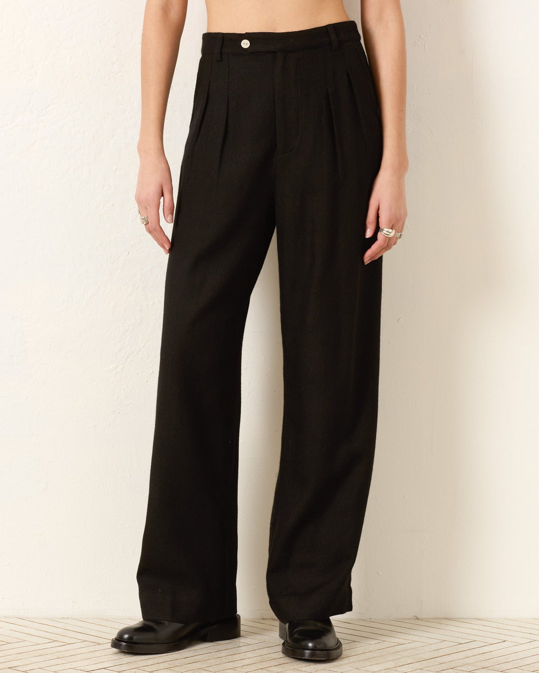 A person wears the Diana Noir Wool Pant—high-waisted, wide-leg black wool with pleats—for a refined menswear-inspired look, paired with black shoes, standing by a light wall. Image shows only the lower body.