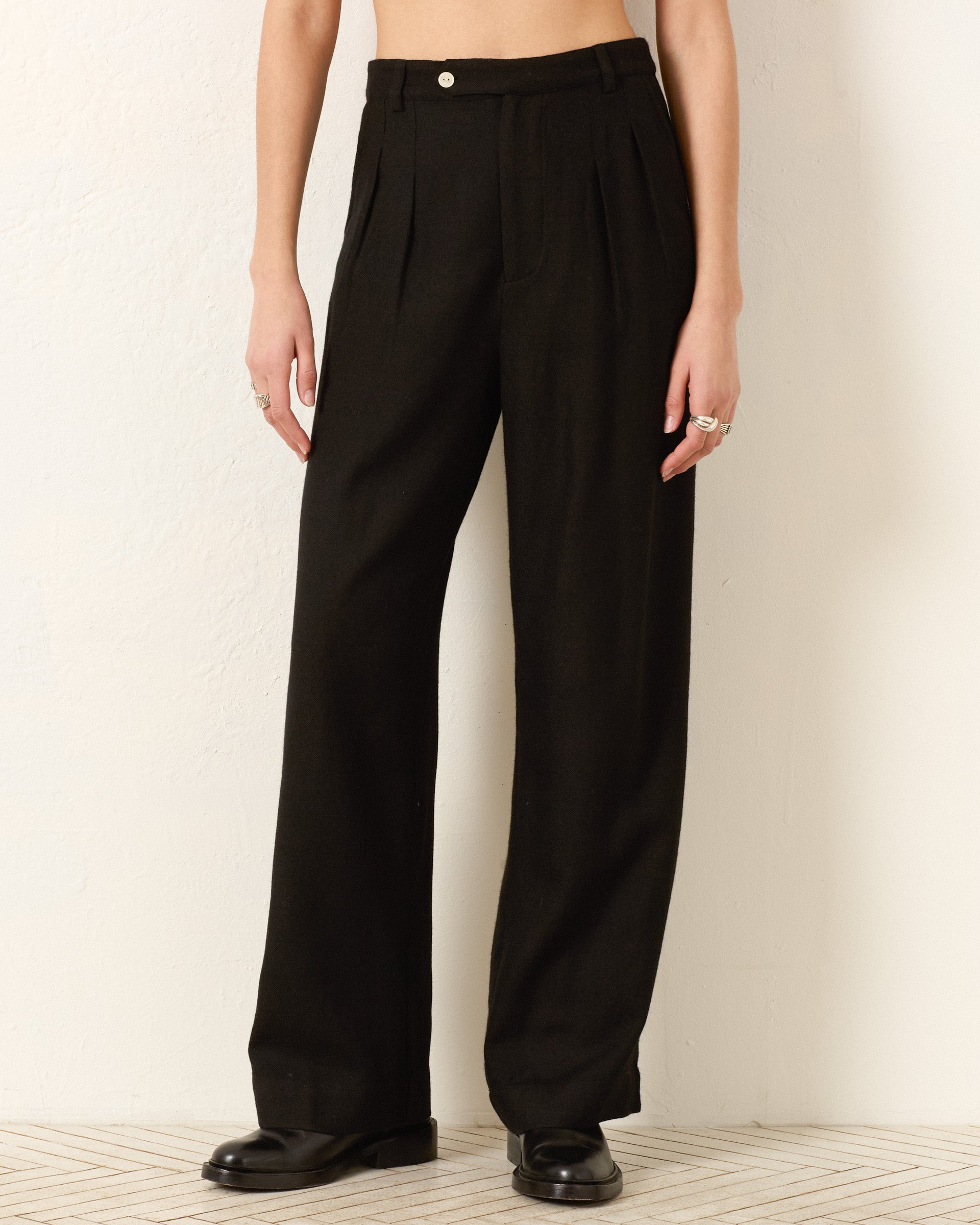 A person wears the Diana Noir Wool Pant—high-waisted, wide-leg black wool with pleats—for a refined menswear-inspired look, paired with black shoes, standing by a light wall. Image shows only the lower body.