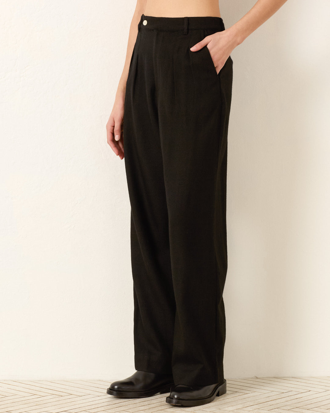 Standing against a light wall, the person wears the Diana Noir Wool Pant—high-waisted black wool pants inspired by menswear tailoring—paired with black shoes and a light-colored top, with one hand in their pocket.