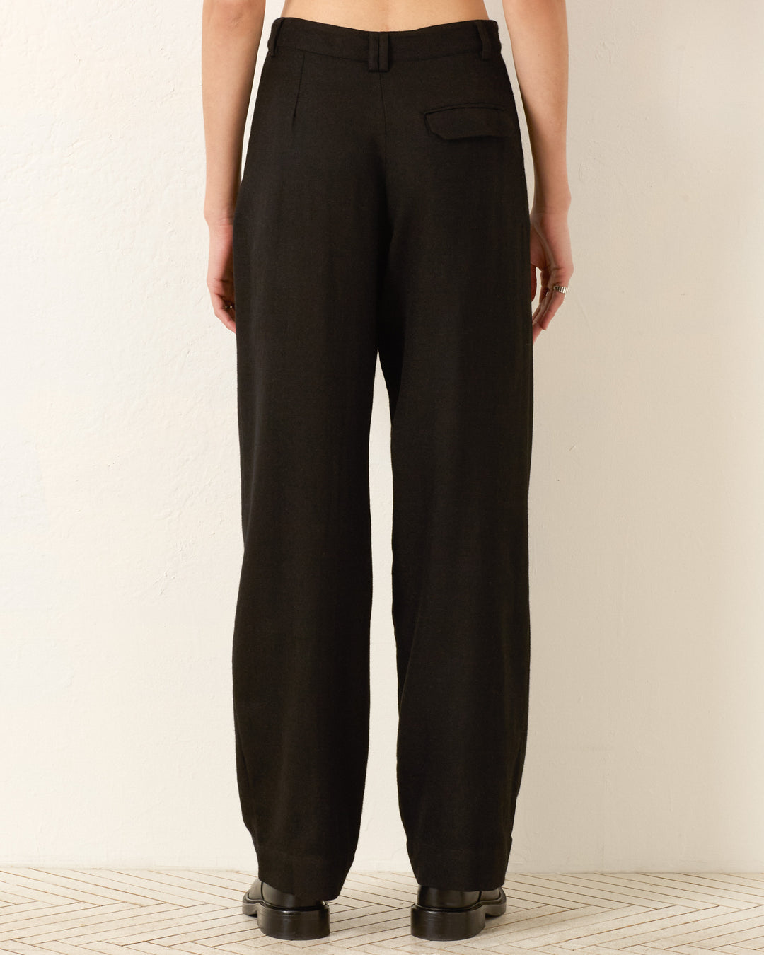 A person stands against a light wall, shown from the back, wearing the Diana Noir Wool Pant—high-waisted wool pants inspired by classic menswear tailoring—and black shoes.