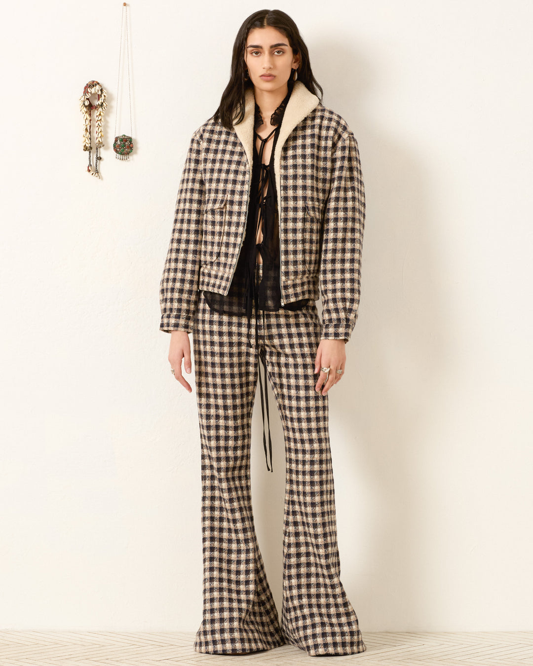 A person stands against a white wall wearing the oversized Eli Trail Dust Bomber Jacket over a black top and flared pants; decorative wall hangings are visible to the left.