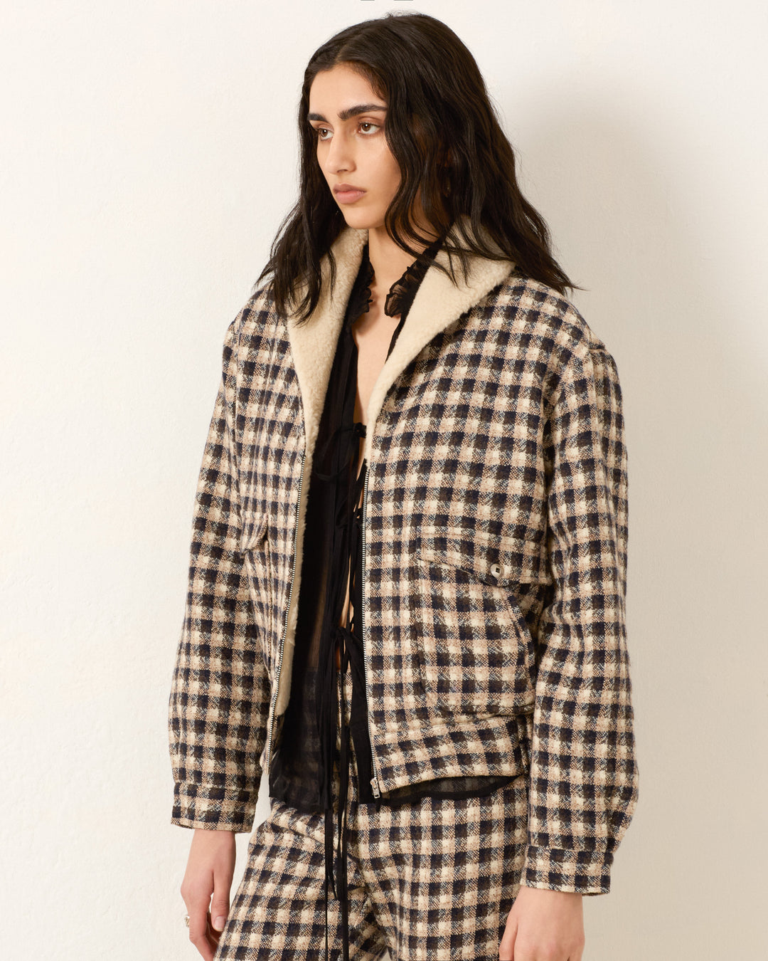 A person with long dark hair wears an oversized-fit Eli Trail Dust Bomber Jacket in black and beige plaid with matching pants, standing against a plain light-colored wall.