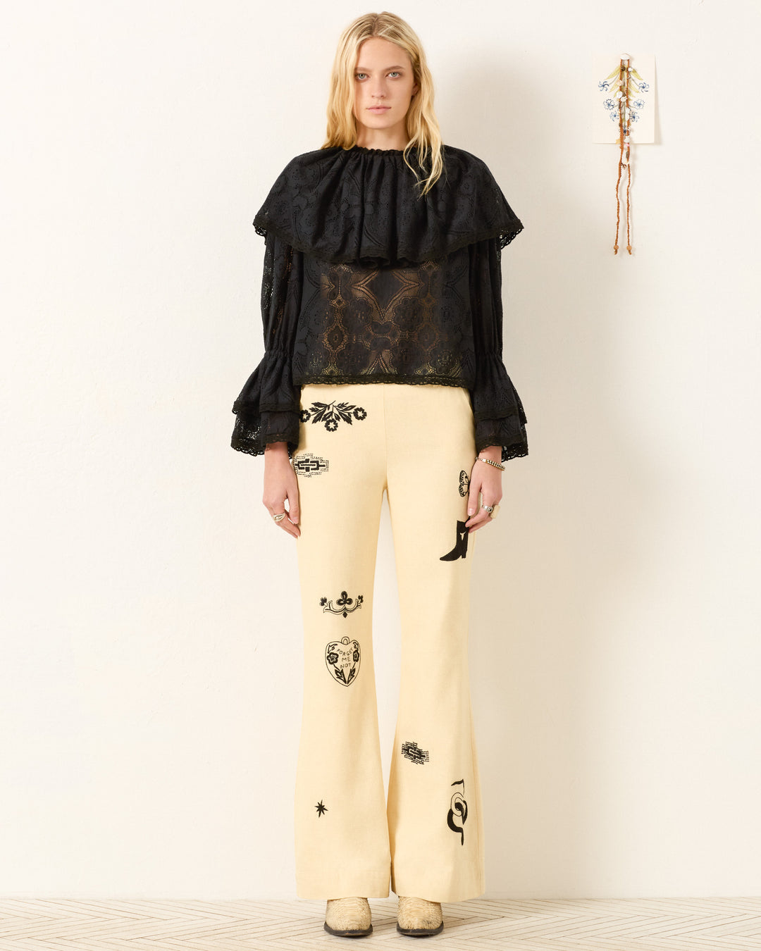 A woman stands against a white wall wearing the Eloise Shadow Lace Top with Victorian sleeves and ruffled collar, paired with cream wide-leg pants featuring black decorative patches.