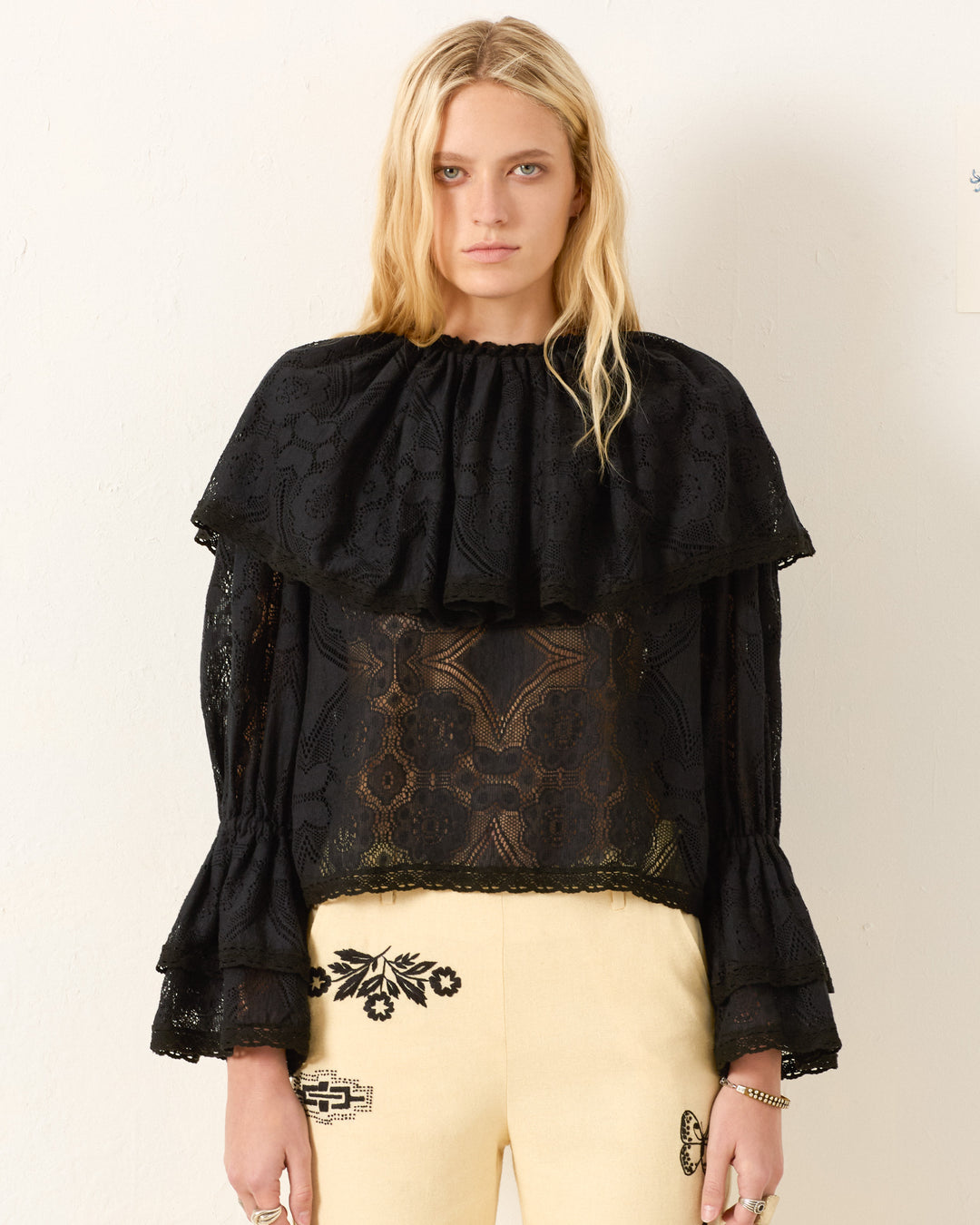 A woman stands against a plain wall wearing the Eloise Shadow Lace Top with Victorian sleeves and a ruffled collar, paired with cream pants featuring black embroidery.