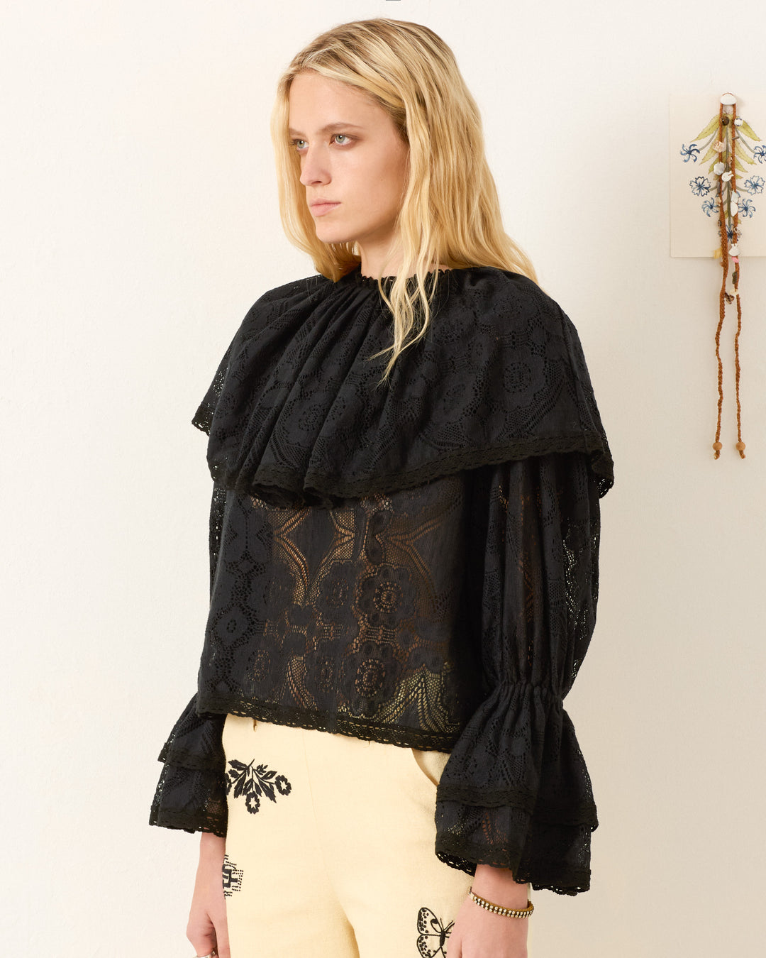 A person with long blonde hair wears the Eloise Shadow Lace Top with Victorian sleeves and cream pants featuring black embroidered designs, standing against a plain wall.