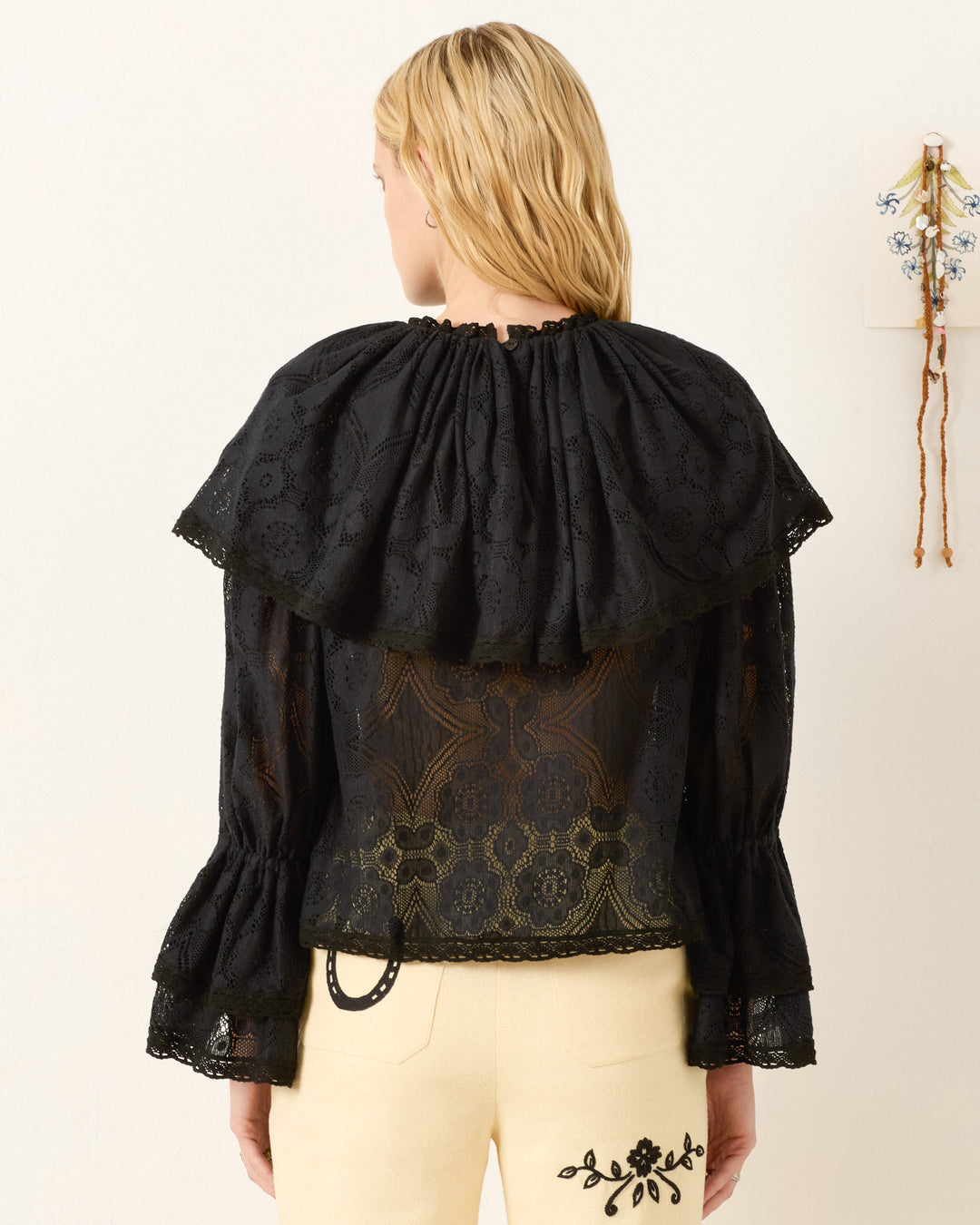 A person with long blonde hair is photographed from the back wearing the Eloise Shadow Lace Top—black floral lace with a wide collar and Victorian sleeves—paired with cream pants featuring black embroidery.