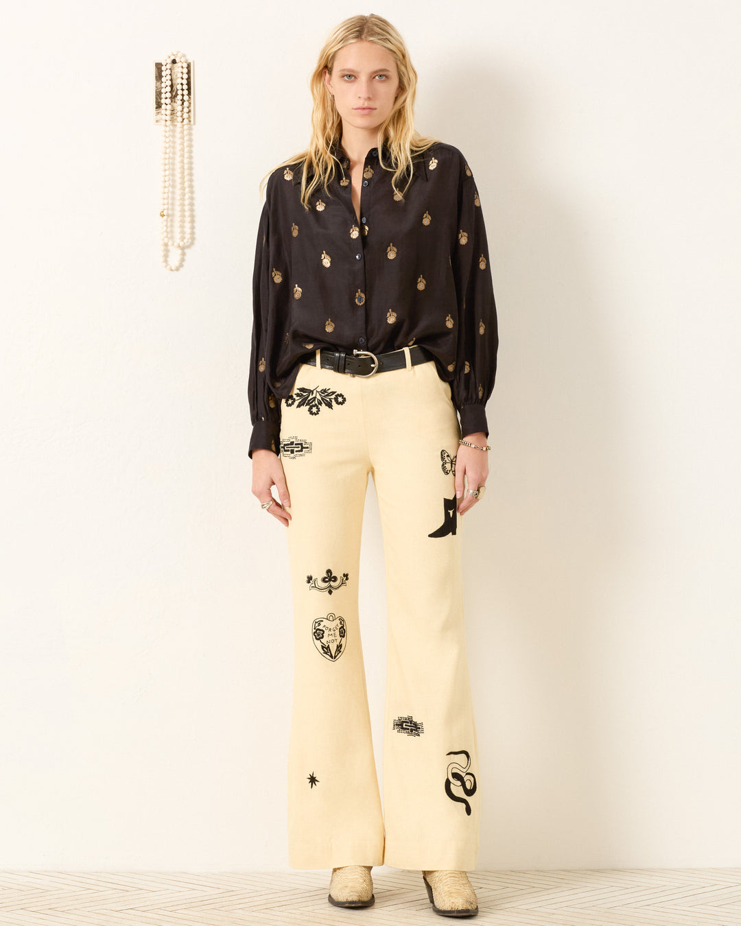 A woman stands against a plain wall wearing the Emery Shadow Bamboo Shirt, cream wide-leg pants with black embroidery, a black belt, and beige shoes. Pearls hang on the wall beside her.