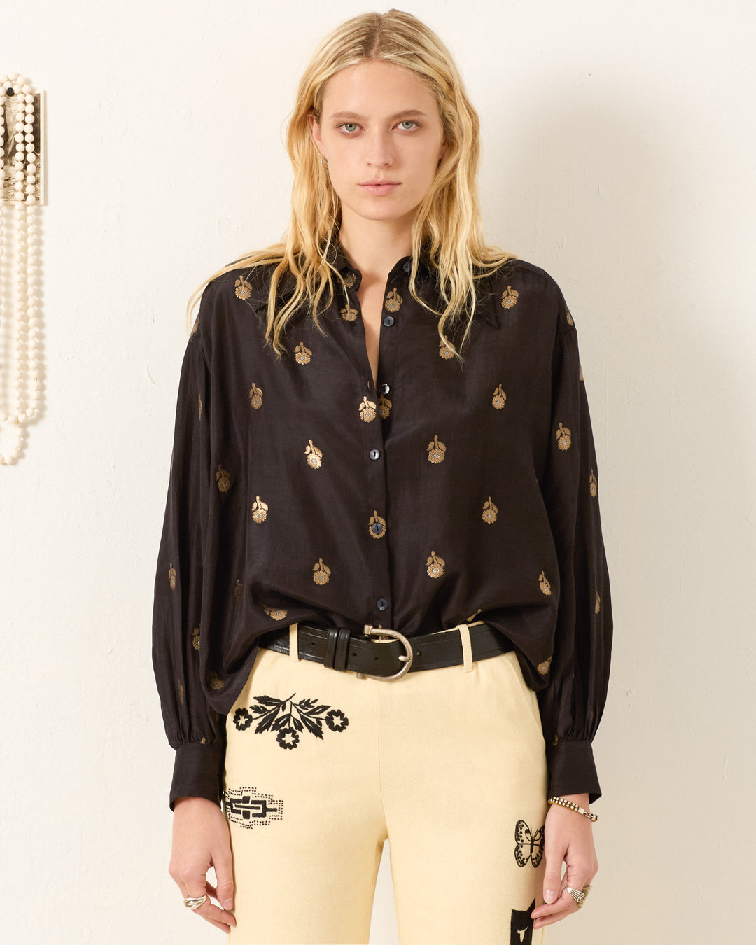 A woman with long blonde hair wears the Emery Shadow Bamboo Shirt, a vintage-inspired black button-up with a metallic gold floral motif, paired with cream pants featuring black embroidery, standing before a plain light background.