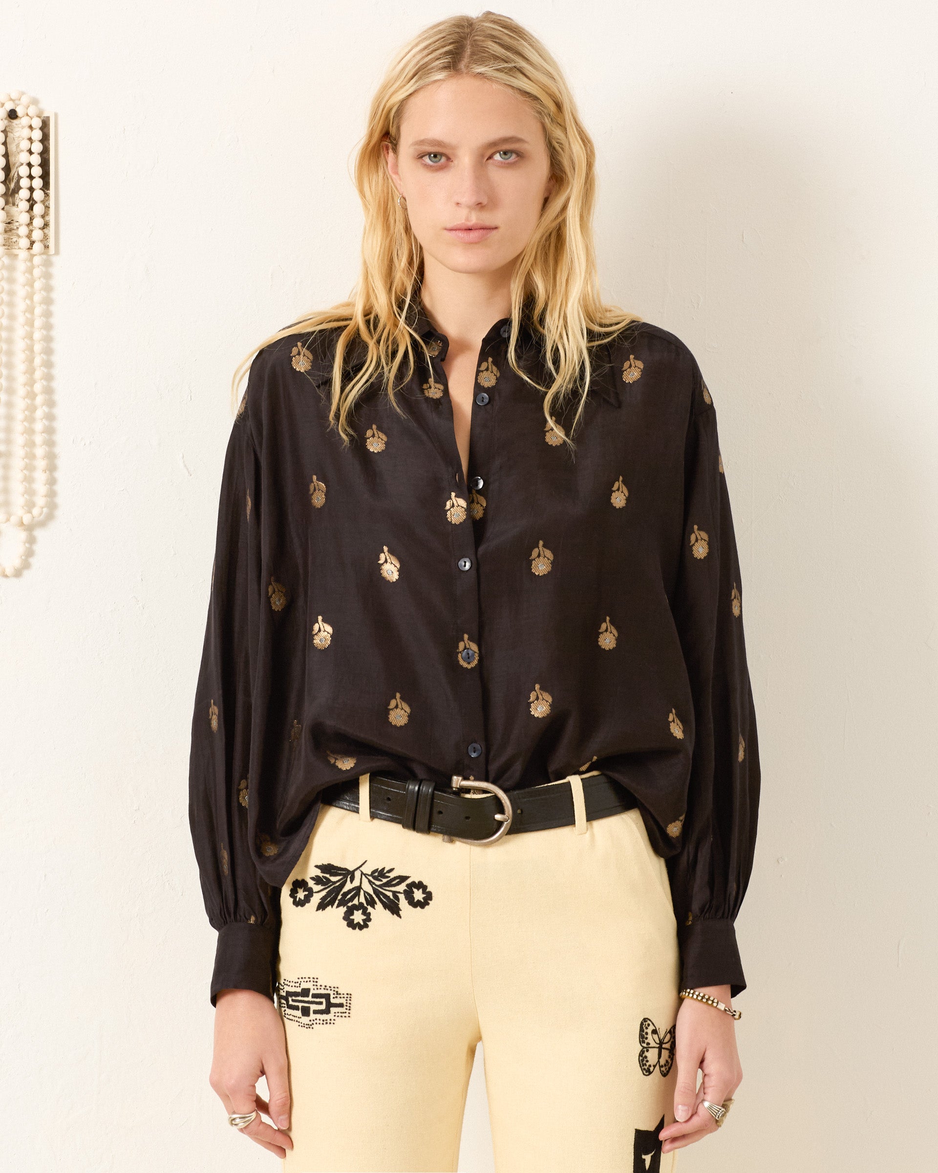 A woman with long blonde hair wears the Emery Shadow Bamboo Shirt, a vintage-inspired black button-up with a metallic gold floral motif, paired with cream pants featuring black embroidery, standing before a plain light background.